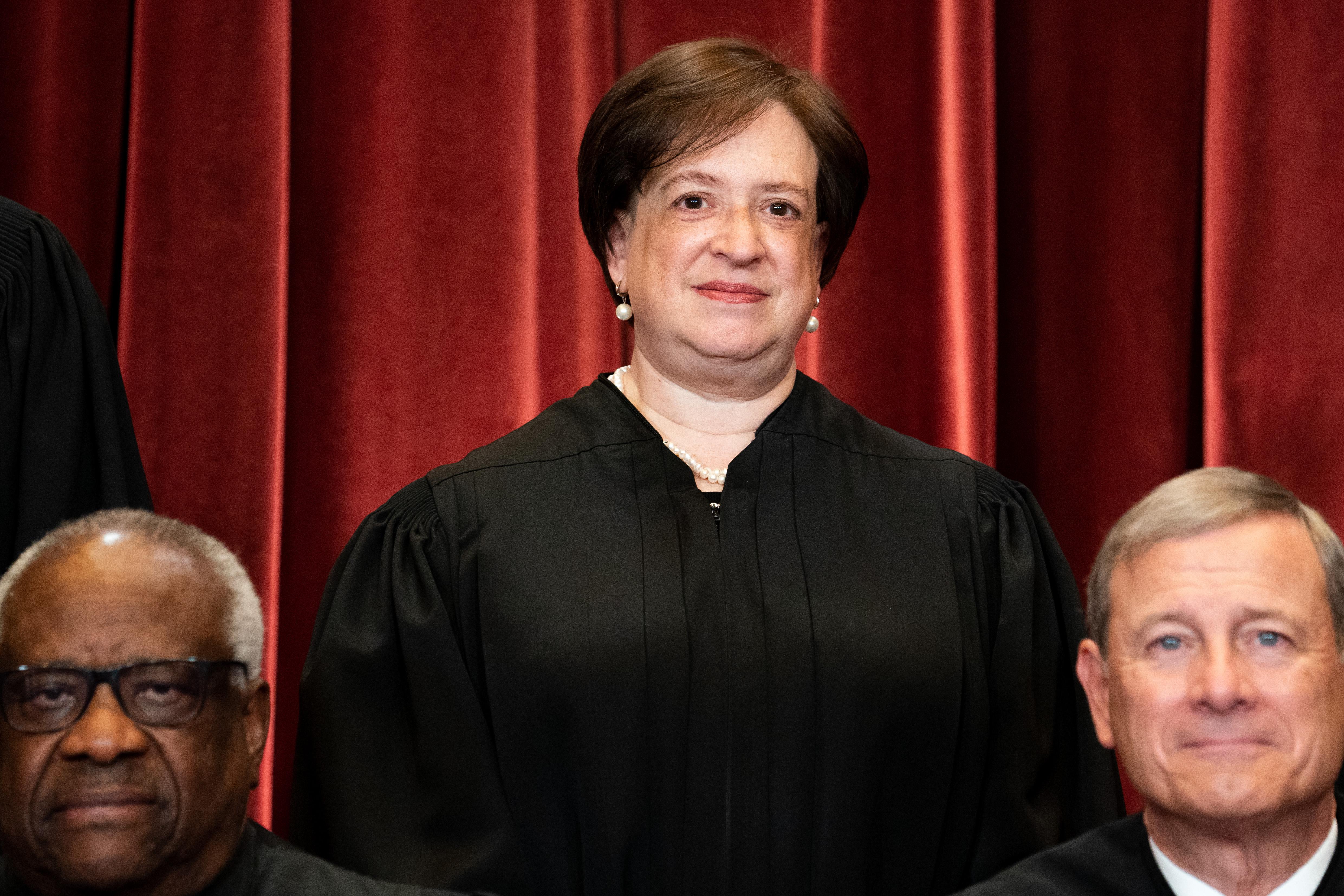 Justice Elena Kagan stands behind Justice Clarence Thomas and Chief Justice John Roberts in a group photo with everyone in their robes in front of a red curtain