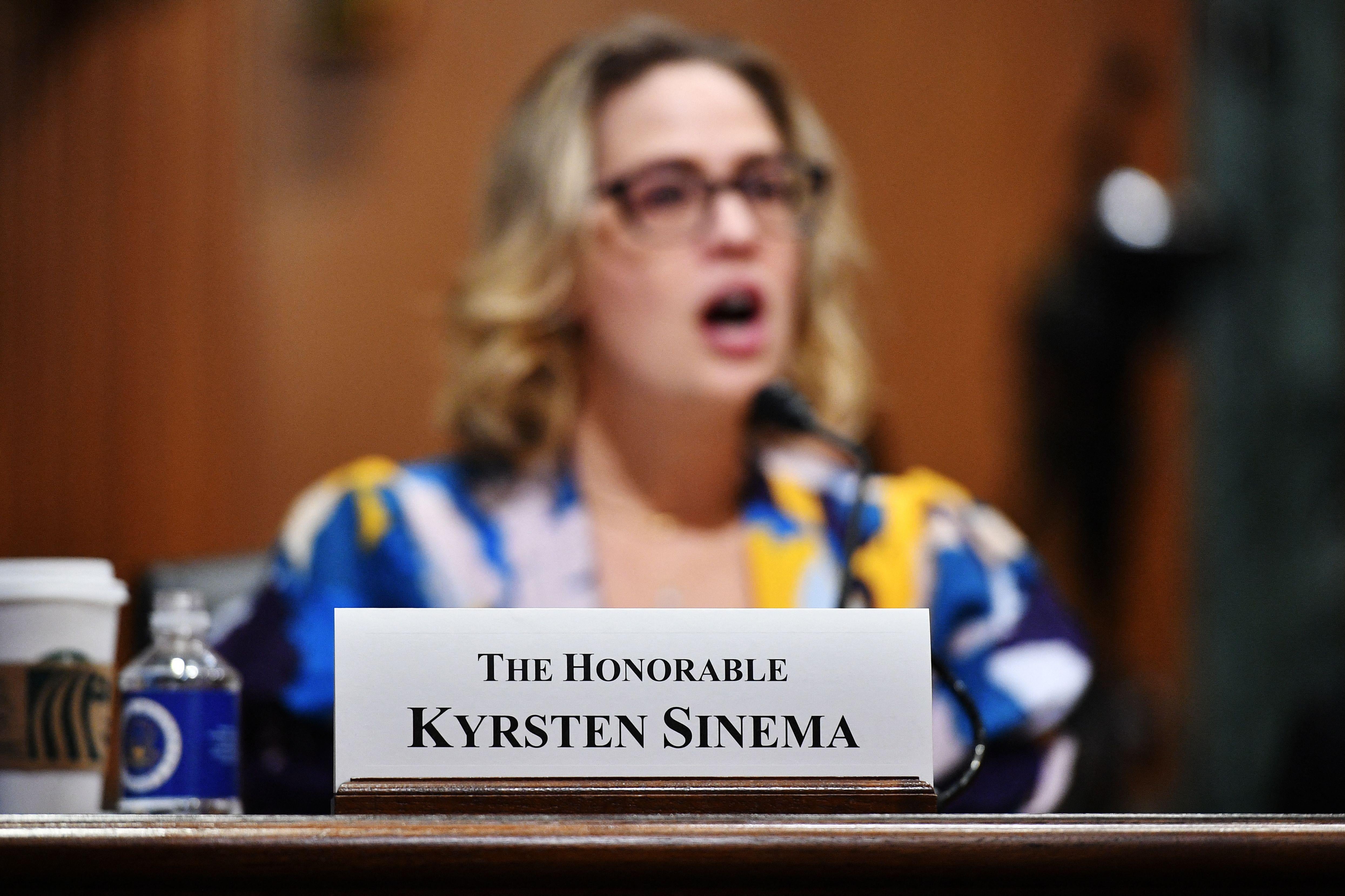Kyrsten Sinema at a hearing, seen behind a sign on the desk that says, "The Honorable Kyrsten Sinema."