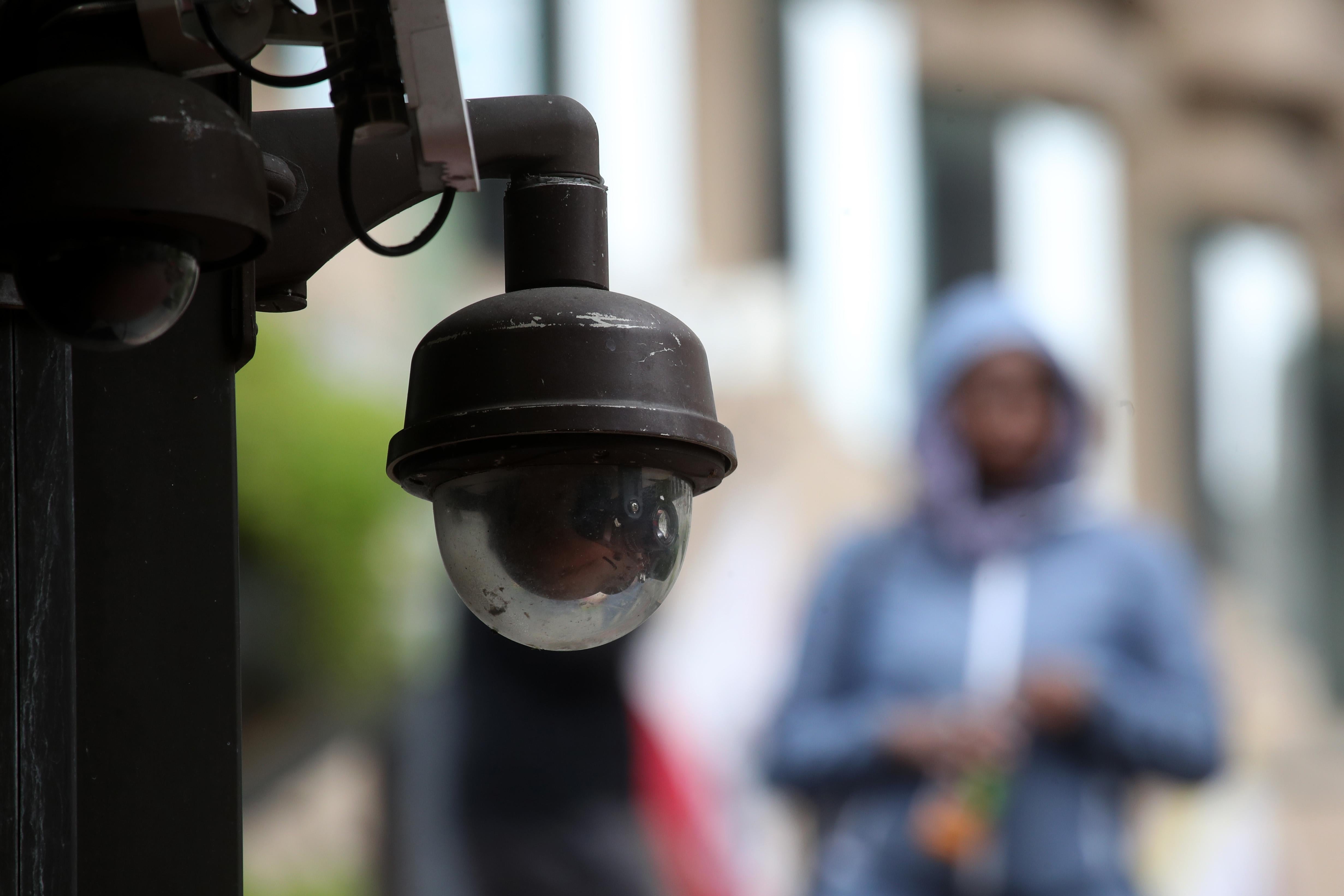 In the foreground, a surveillance camera; in the background, a person wearing a hoodie.