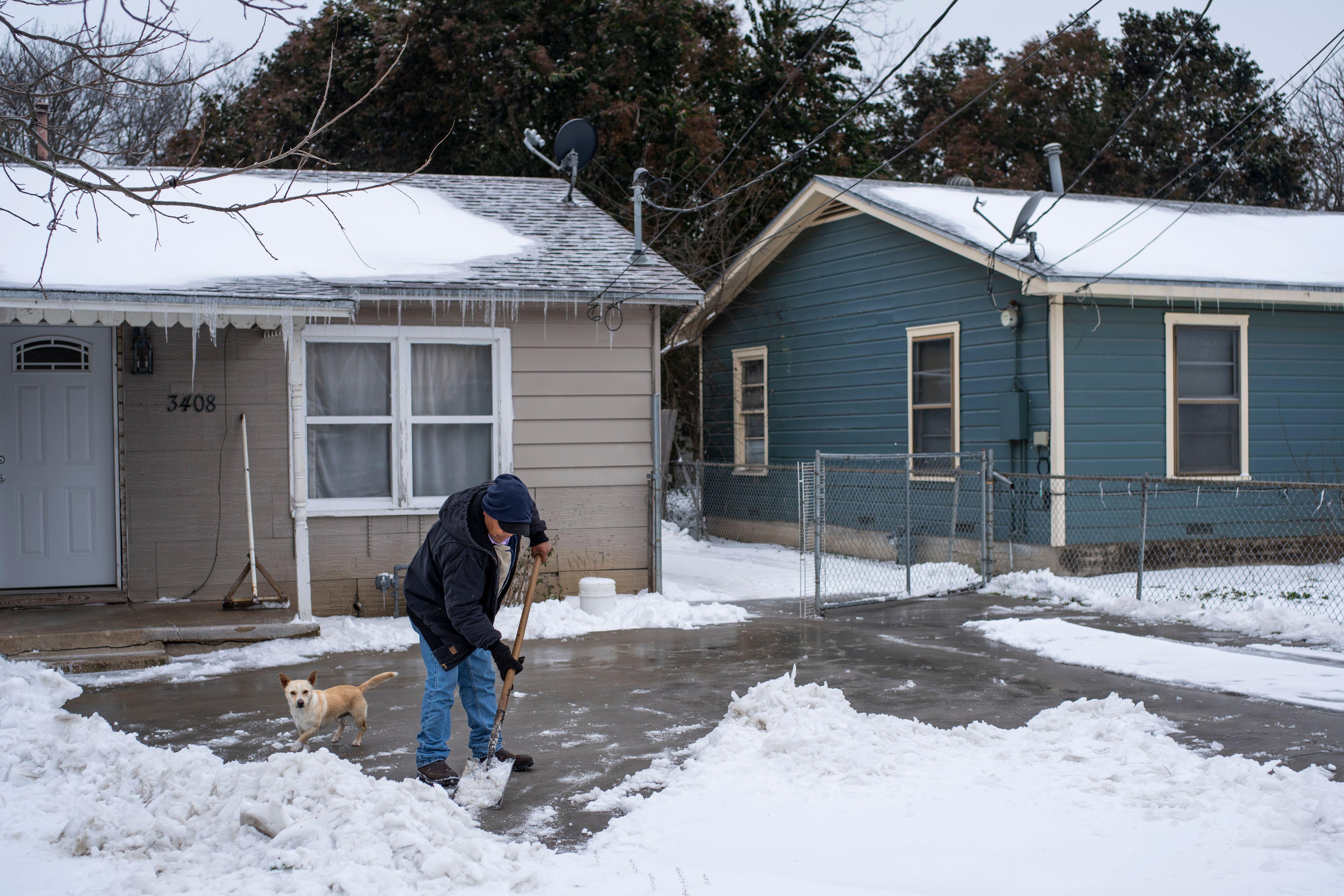 A man shovels snow from his driveway as his dog stands nearby.
