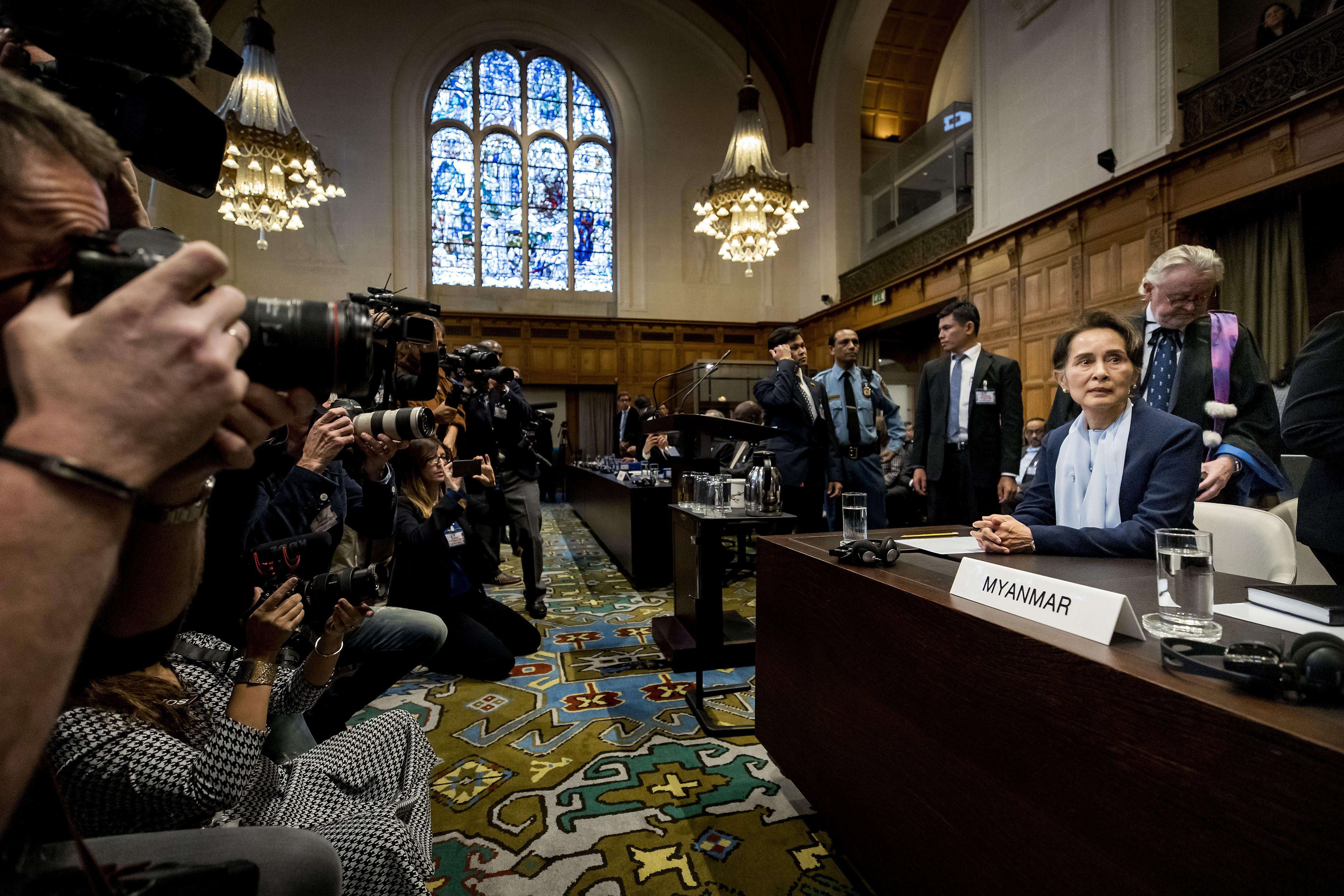 Aung San Suu Kyi sits behind a placard marked "Myanmar" in a courtroom. Photographers to her left hold up cameras to take pictures of her.