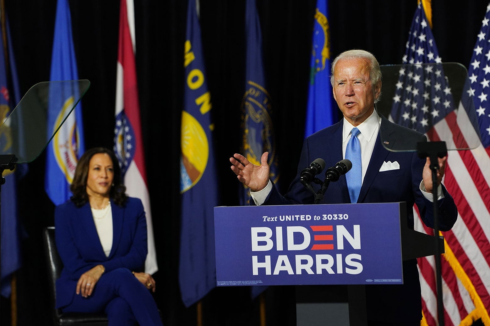 Biden gestures with his arms while speaking at a podium. Harris is seated behind him onstage.