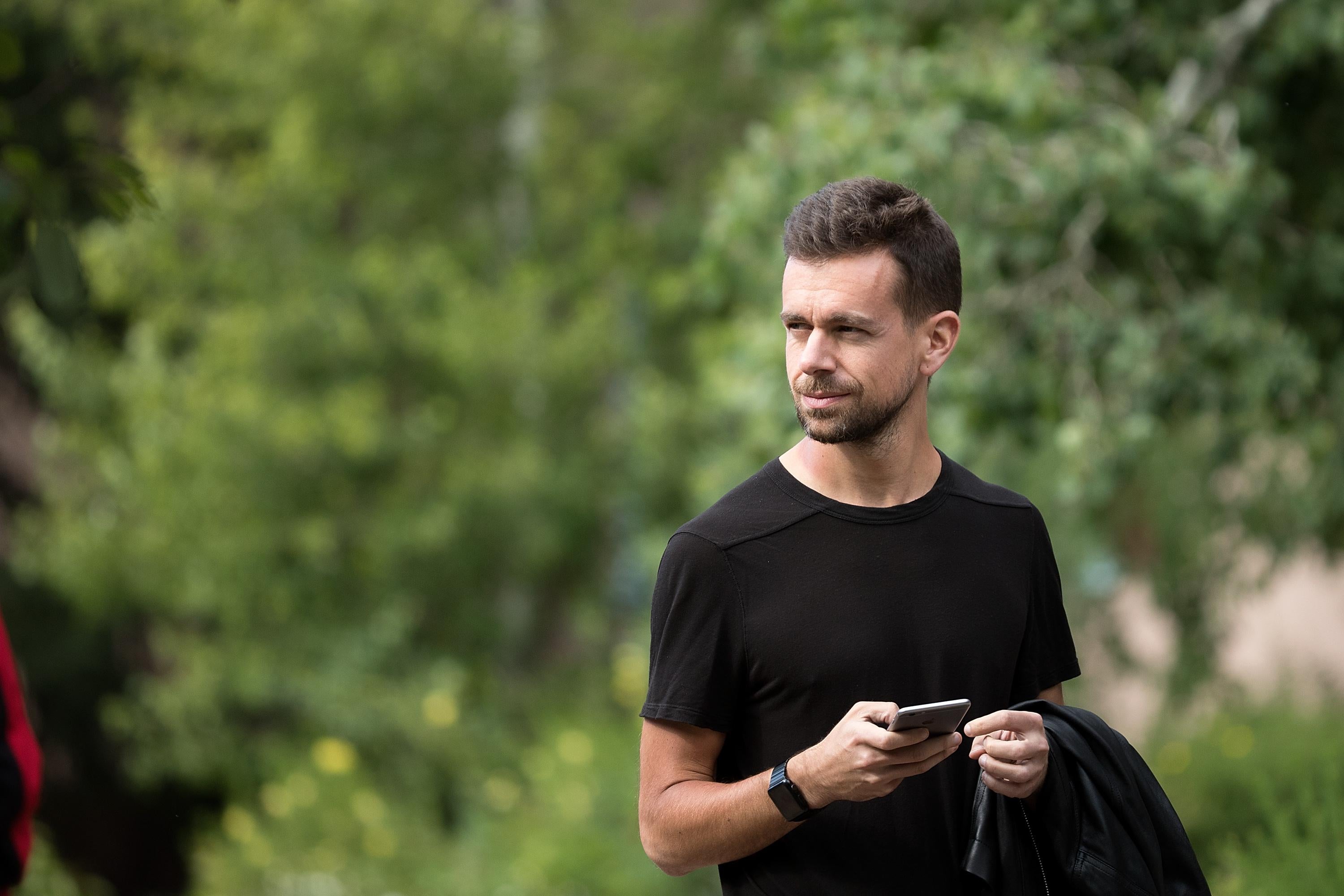 Jack Dorsey in front of some greenery, wearing all black, and holding his mobile phone.