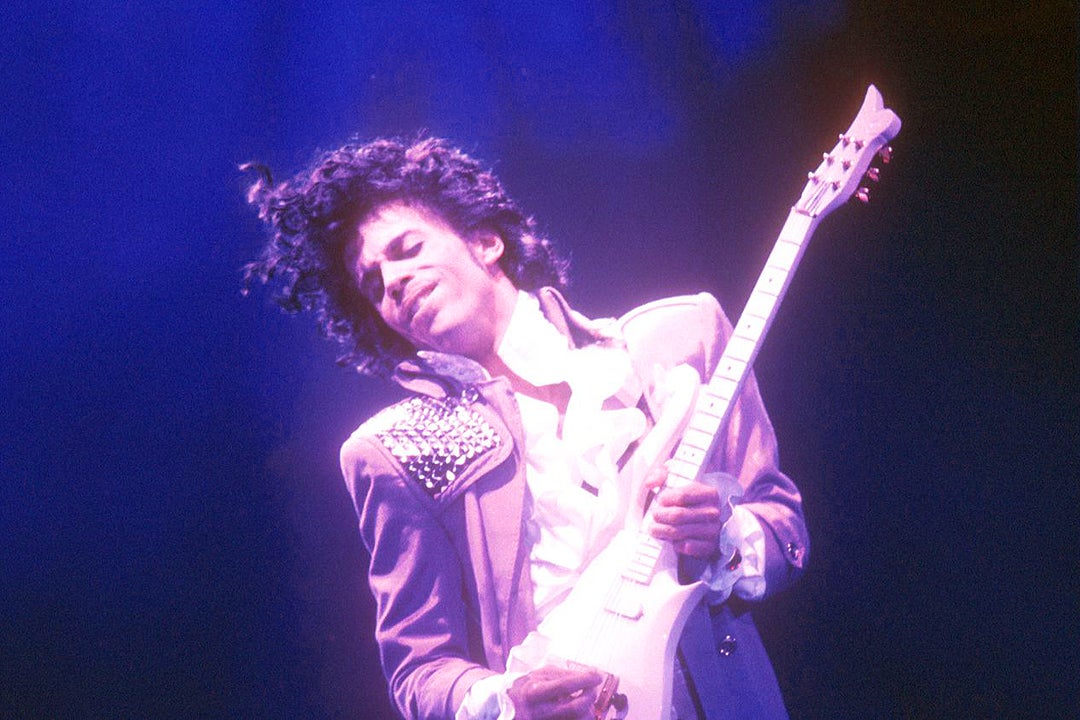 Prince, bathed in purple light, holds a guitar.