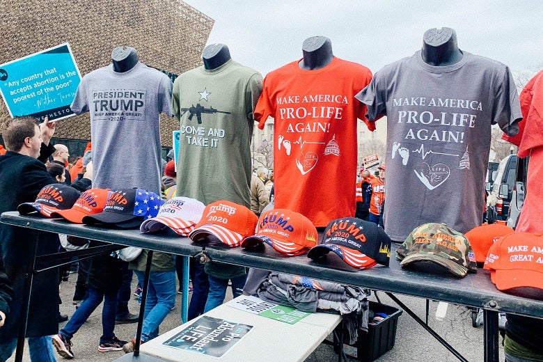 People march behind a T-shirt display with four shirts displayed. One says "President Trump Keep America Great." One has a rifle on it and says "Come and Take It." The other two say "Make America Pro-Life Again."