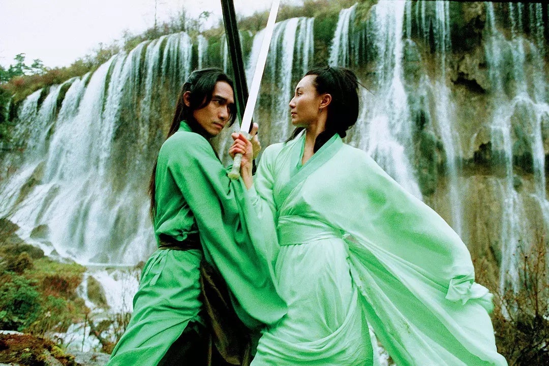 In front of a waterfall, a man and woman, both wearing green, cross swords.