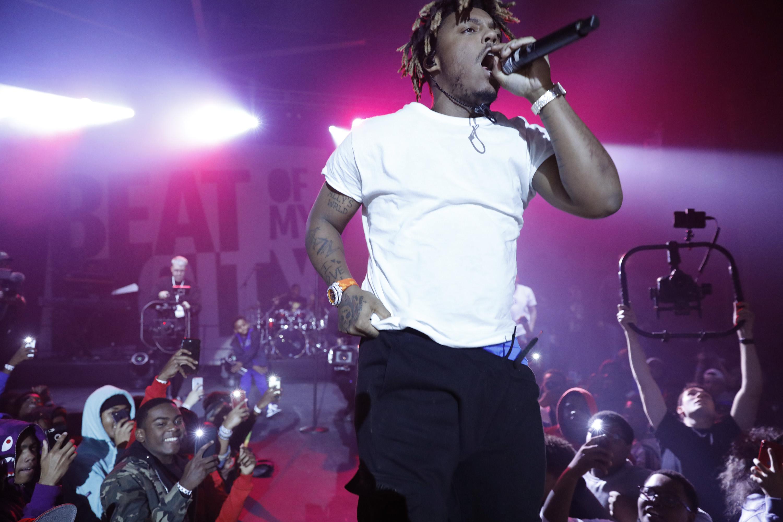 Juice Wrld, wearing a white t-shirt, black pants, and an orange watch, holding a microphone to his mouth on stage.
