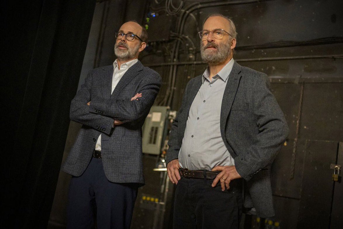 Two bearded, bespectacled middle-aged men in navy suits stare into the distance.