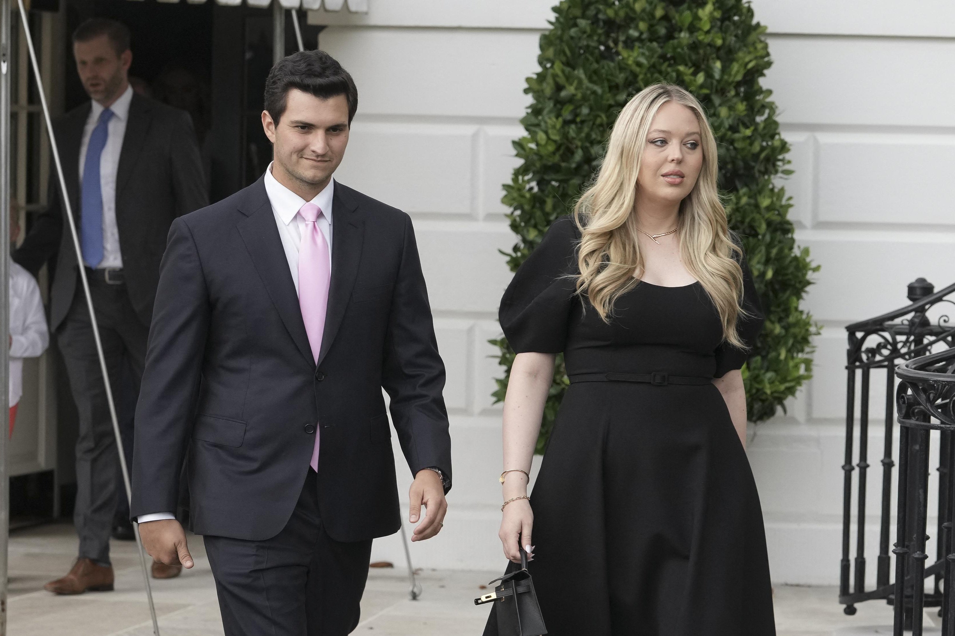 Tiffany Trump wears black and walks alongside her husband, in a black suit, in a tiled White House courtyard with a party tent in it. 