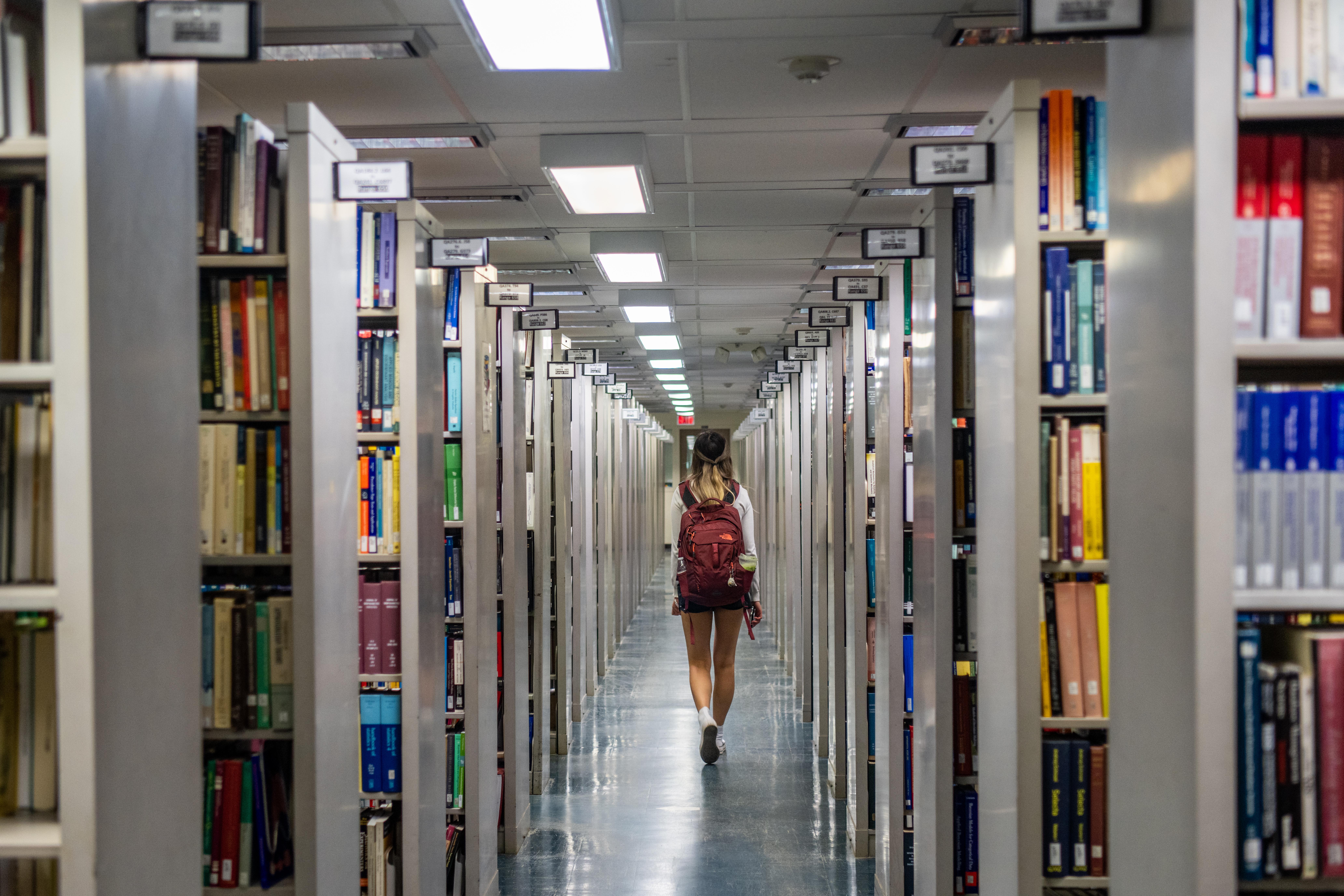 A person with a backpack walks through tall library shelves filled with books.