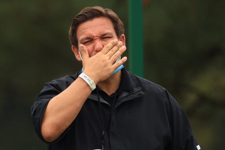 DeSantis with a surgical mask over his chin holds his hand over his mouth as he yawns outside