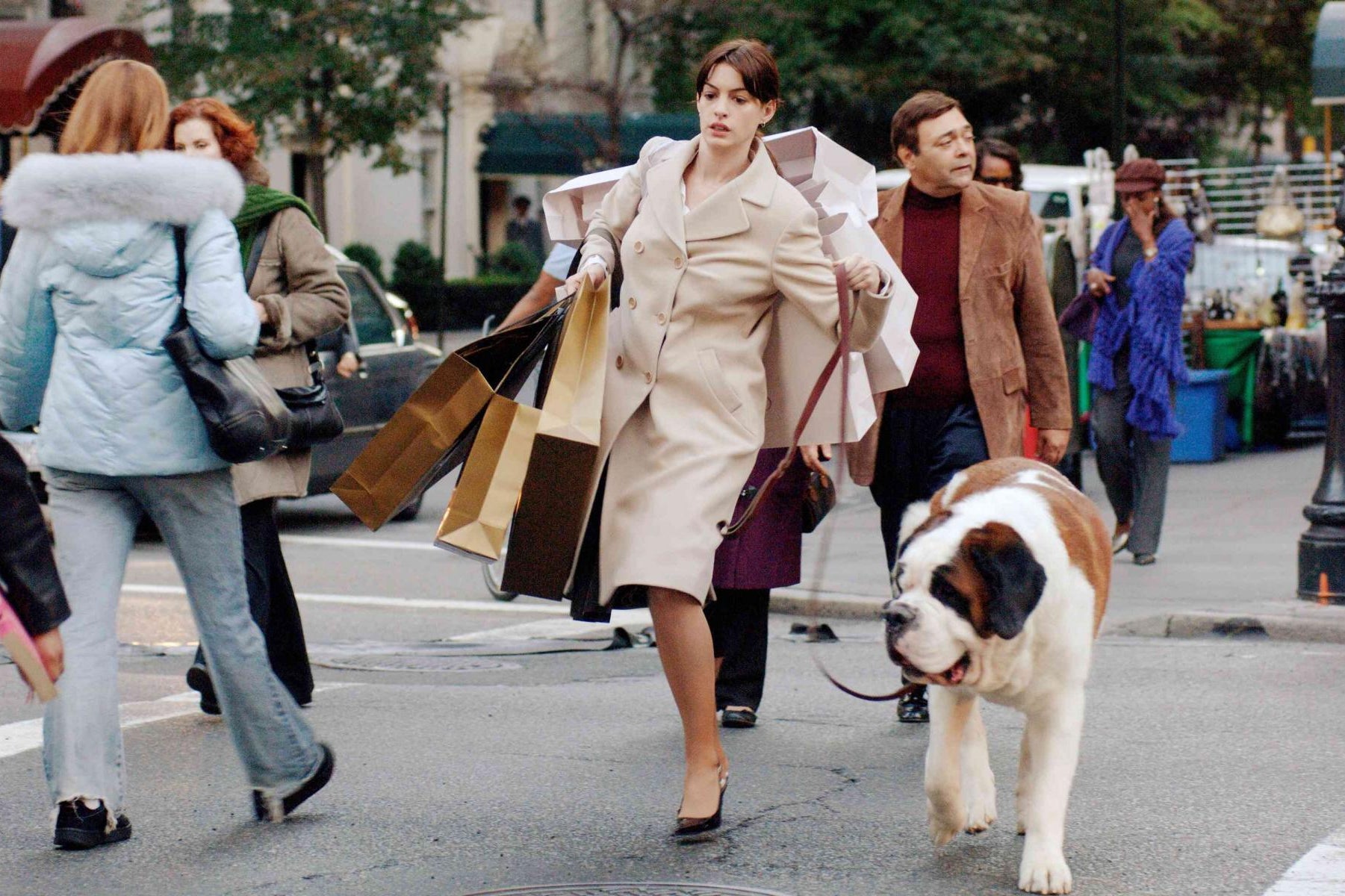 A white woman with brown hair, pulled back in a low ponytail, wearing a two-piece white suit/skirt set and heels, is rushing across a busy New York crosswalk. She is holding multiple shopping bags in both of her hands, as well as has her purple purse hanging from her arm. In her left hand, she is also holding a leash and walking a large Saint Bernard dog. 