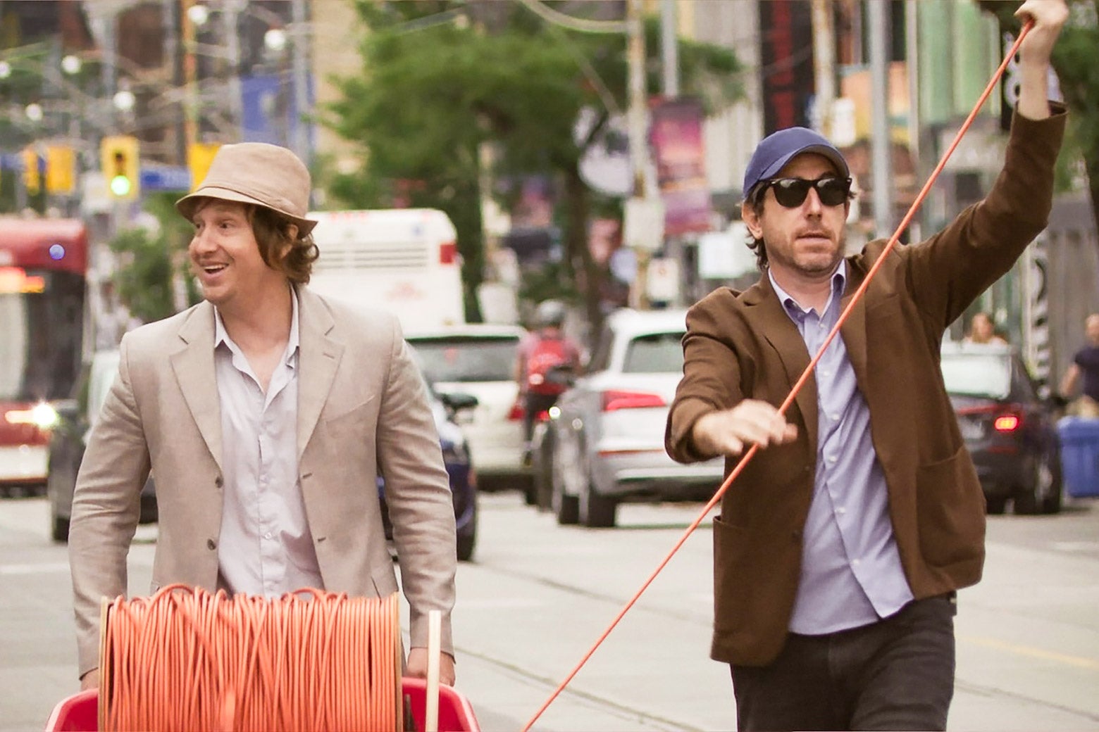 A white man wearing a beige suit and fedora smiles and walks down the street, while a white man in a blue baseball hat walks next to him, unraveling an orange cord.