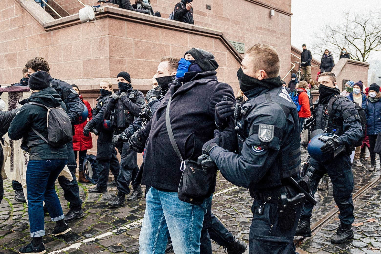 Police stand between Querdenken supporters and left-wing counterprotesters. In the foreground, two officers hold a man by both arms.