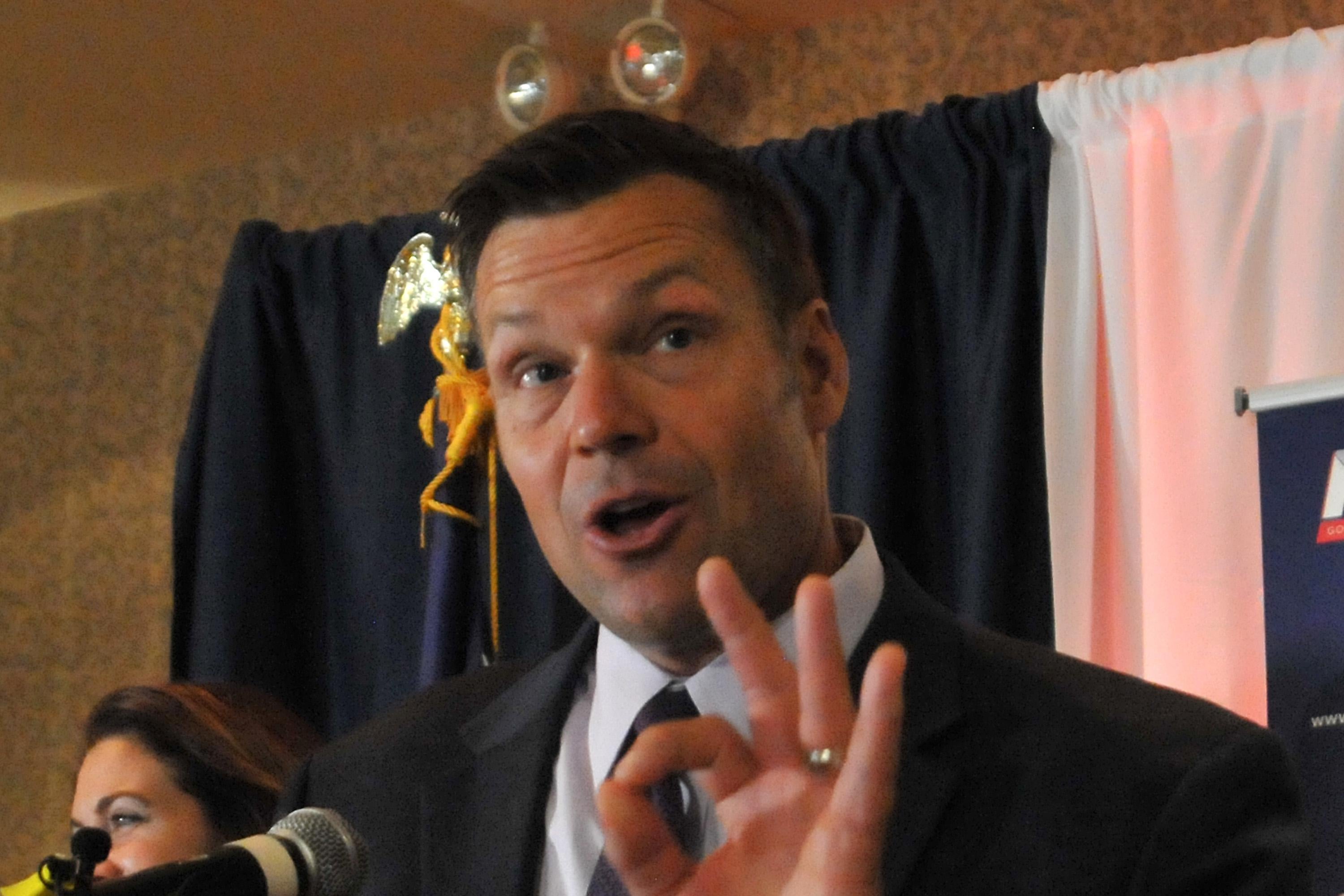 Kris Kobach speaks at a microphone and makes a gesture with his hand.