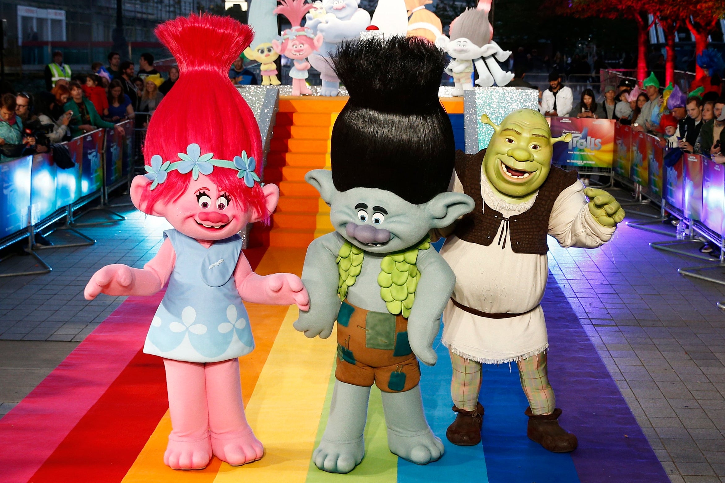 Two people dressed in mascot costumes as trolls from the animated movie Trolls, plus one person dressed as Shrek, walking down a rainbow carpet at a Trolls photocall.
