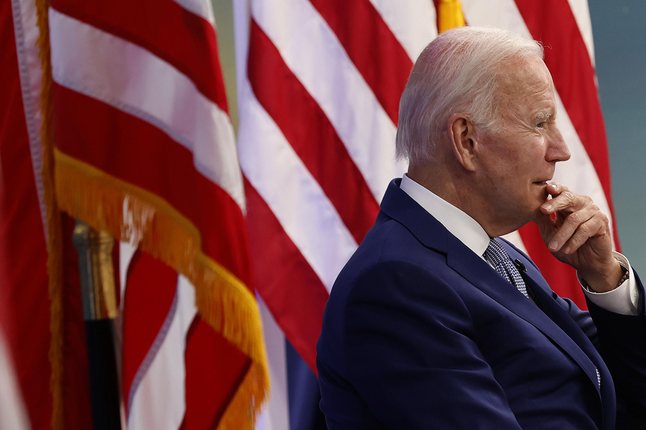 A photo of President Joe Biden, from the side, in front of an American flag.