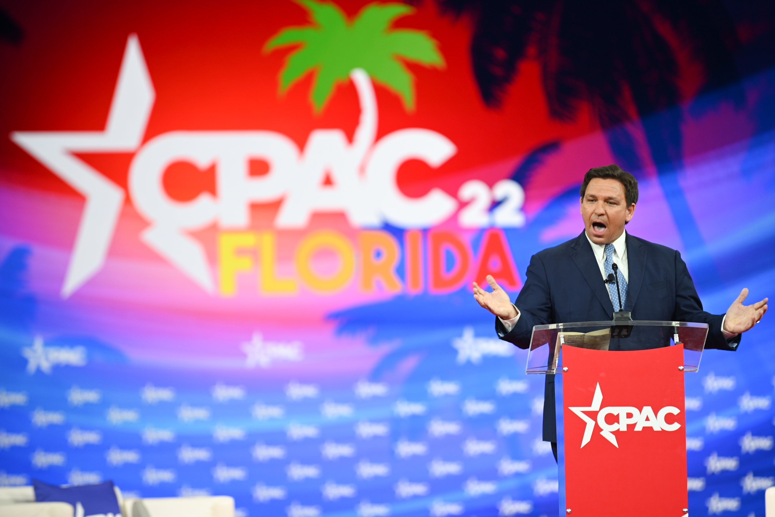 DeSantis gestures as he speaks at a lectern with a tropical CPAC backdrop behind him