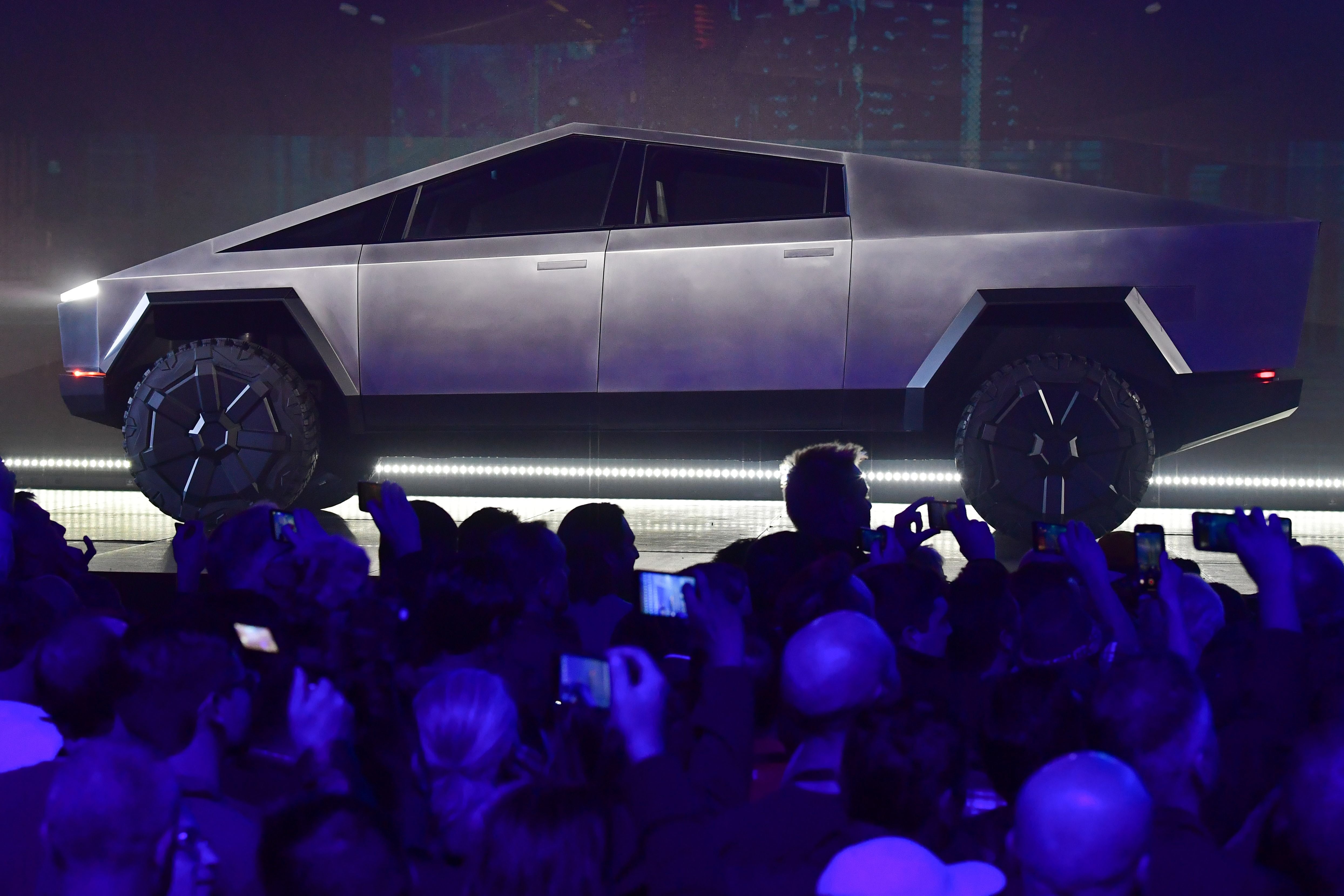 The very angular and matte-gray Cybertruck is on display on a stage above a crowd of people's heads at the Tesla Design Center in California. Almost everyone has their phone raised to take a picture. 