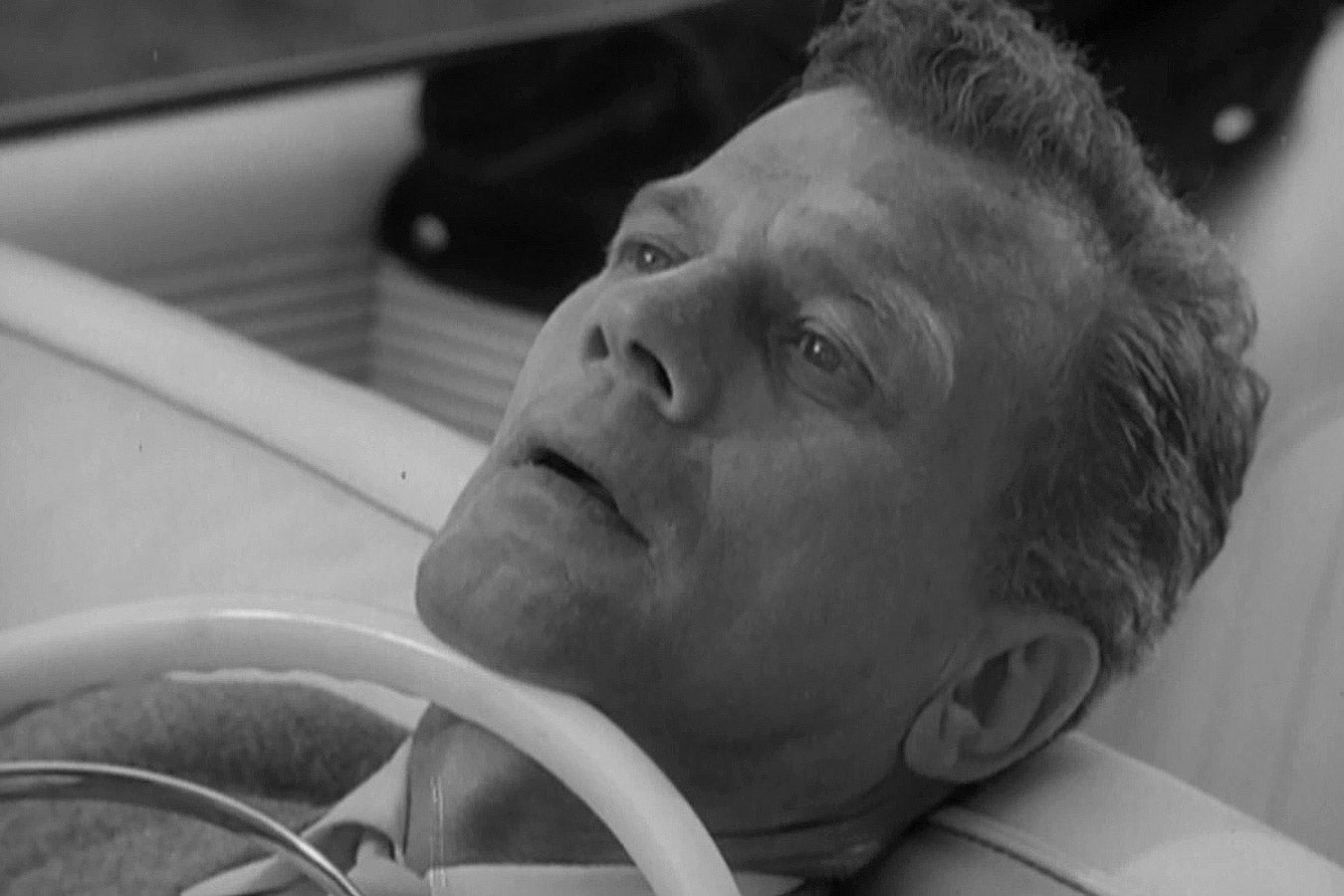 A man lies in a car with the steering wheel pressing into his neck.