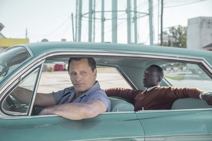 Two men in a light, blue-ish green car. One man is sitting in the front seat with his elbow out the window. The other is in the backseat, arms stretched across.