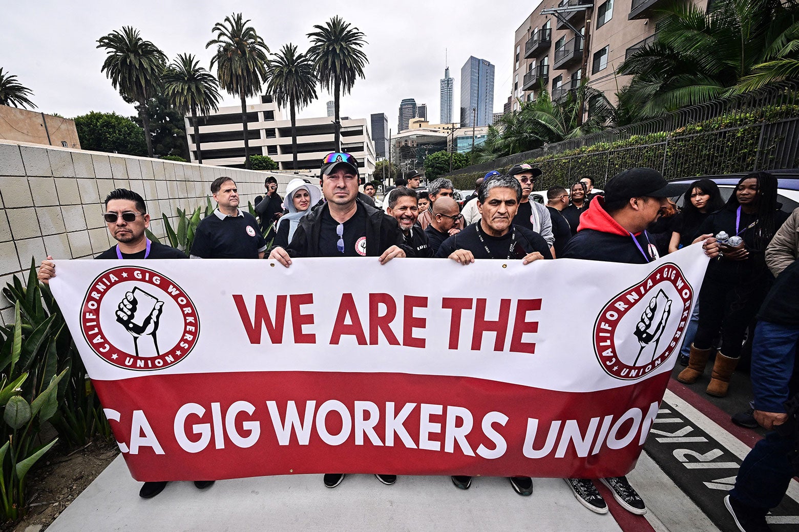 A large group marches behind a banner that says "We are the CA Gig Workers Union"; palm trees and a downtown skyline can be seen behind them.