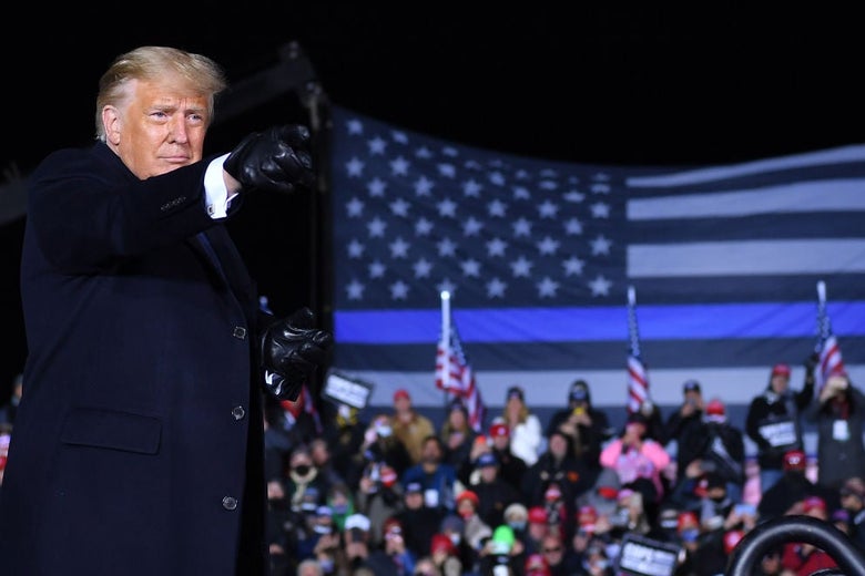 Trump points with a gloved hand, a large Thin Blue Line flag and bleachers full of supporters behind him.