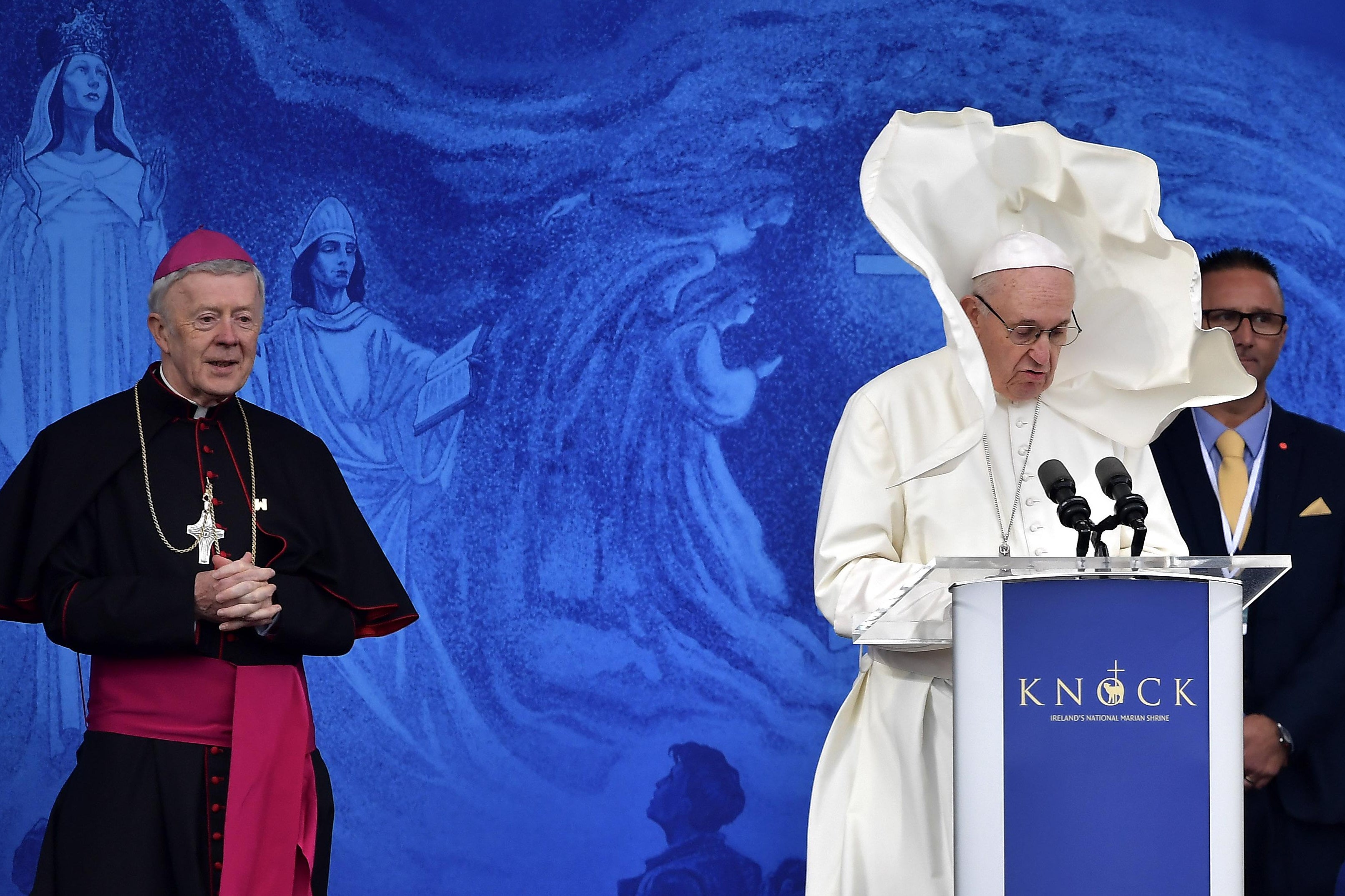 Pope Francis' cape blows straight up in the air as he speaks at a podium.