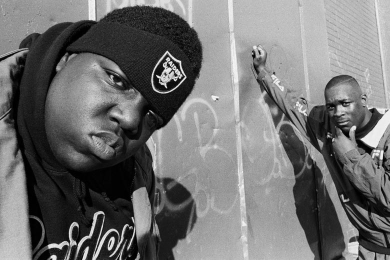 The rapper scowls at the camera in a Raiders headband while another man poses against a wall in the background.