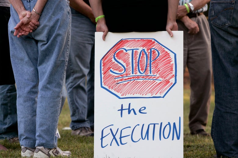 Person holding a sign that says "Stop the Execution" among a group of people seen at waist level standing on grass