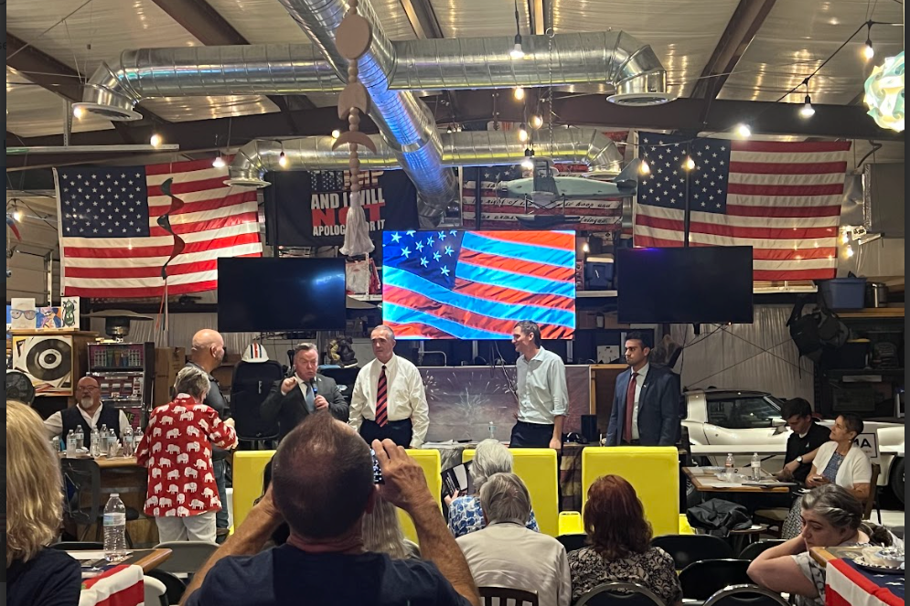 Candidates speak to a crowd in front of several American flags inside a garage.