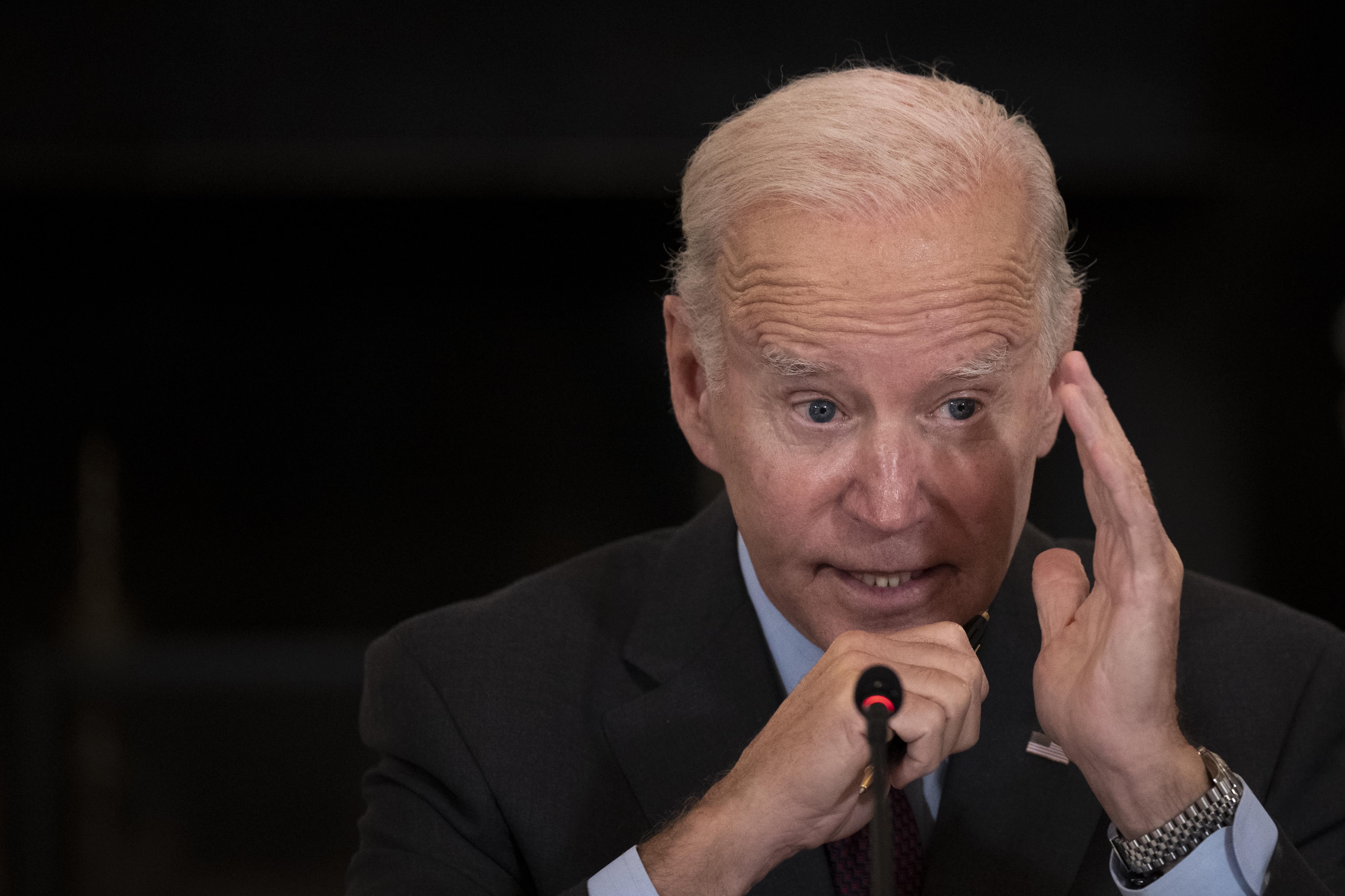 Biden speaking into a microphone with his left hand raised next to his face