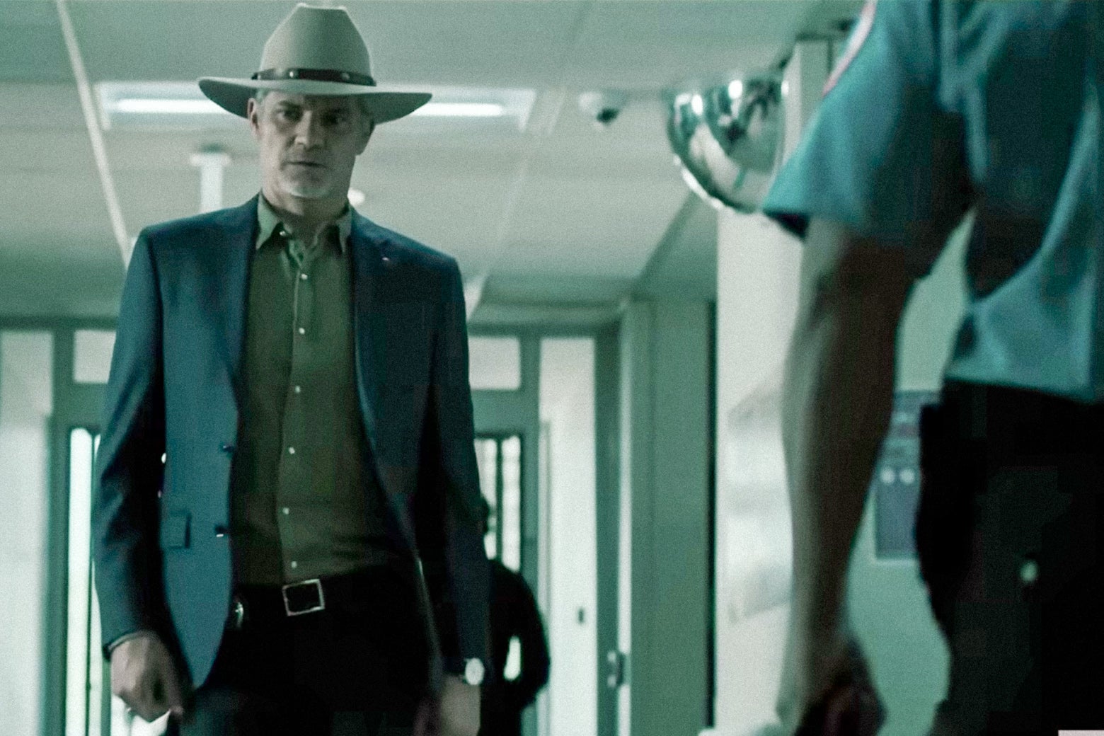 A defeated-looking man in a cowboy hat walks down a green-toned hallway, looking downward.