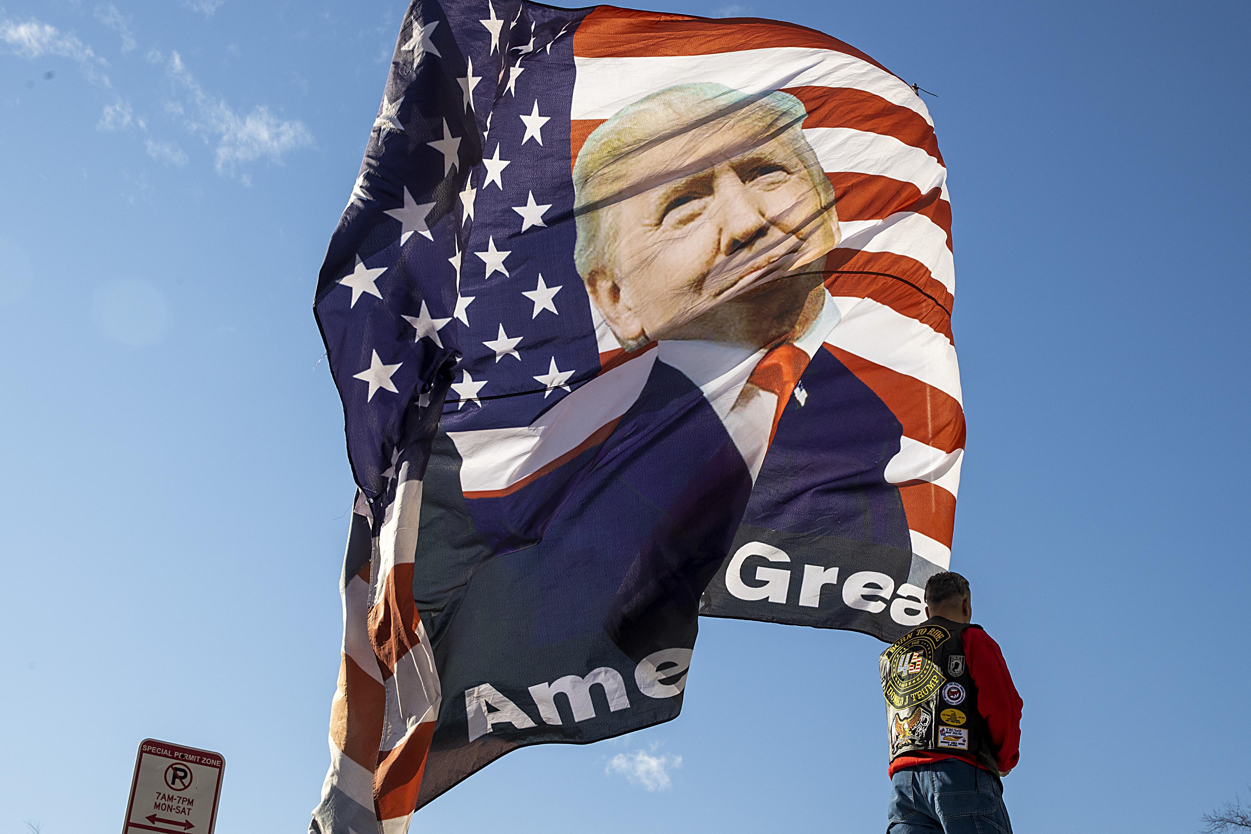 Person standing under a giant MAGA flag with Trump's face on it, waving against a blue sky in Washington