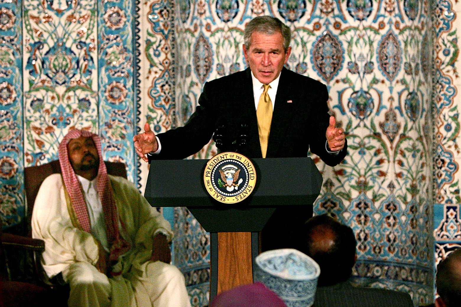 Bush gestures with his hands while speaking at a lectern to a group of Muslims. A Muslim leader is seated to his right.