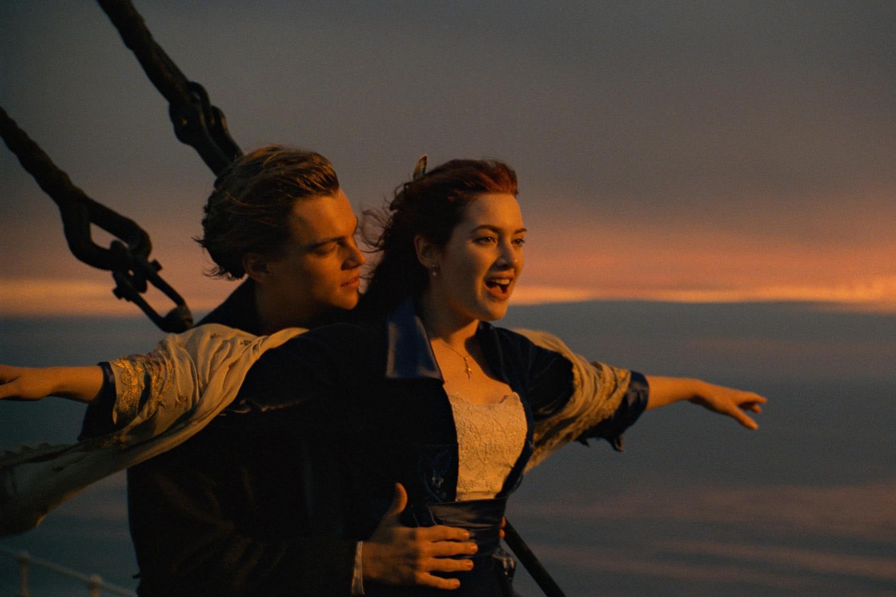 At sunset, Leonardo DiCaprio as Jack holds the waist of Kate Winslet as Rose as she stands at the front of a ship with her arms out at her sides and an exhilarated expression on her face.