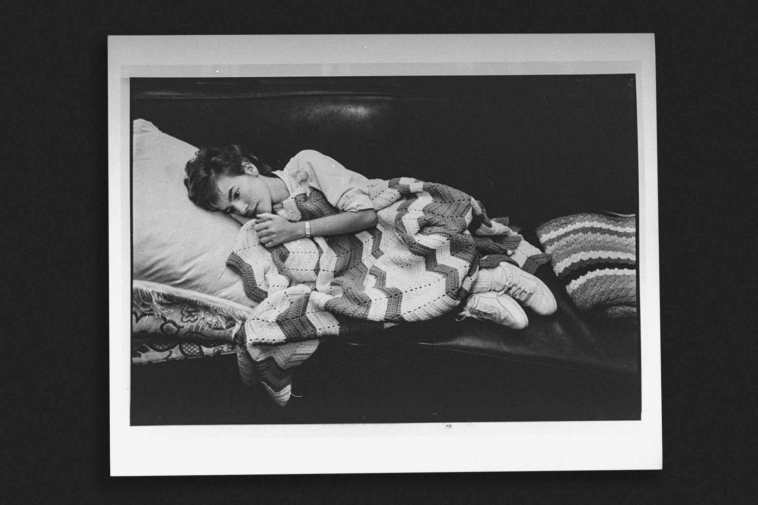 Black-and-white photo of a young woman with short blond hair lying on a couch covered by a blanket.