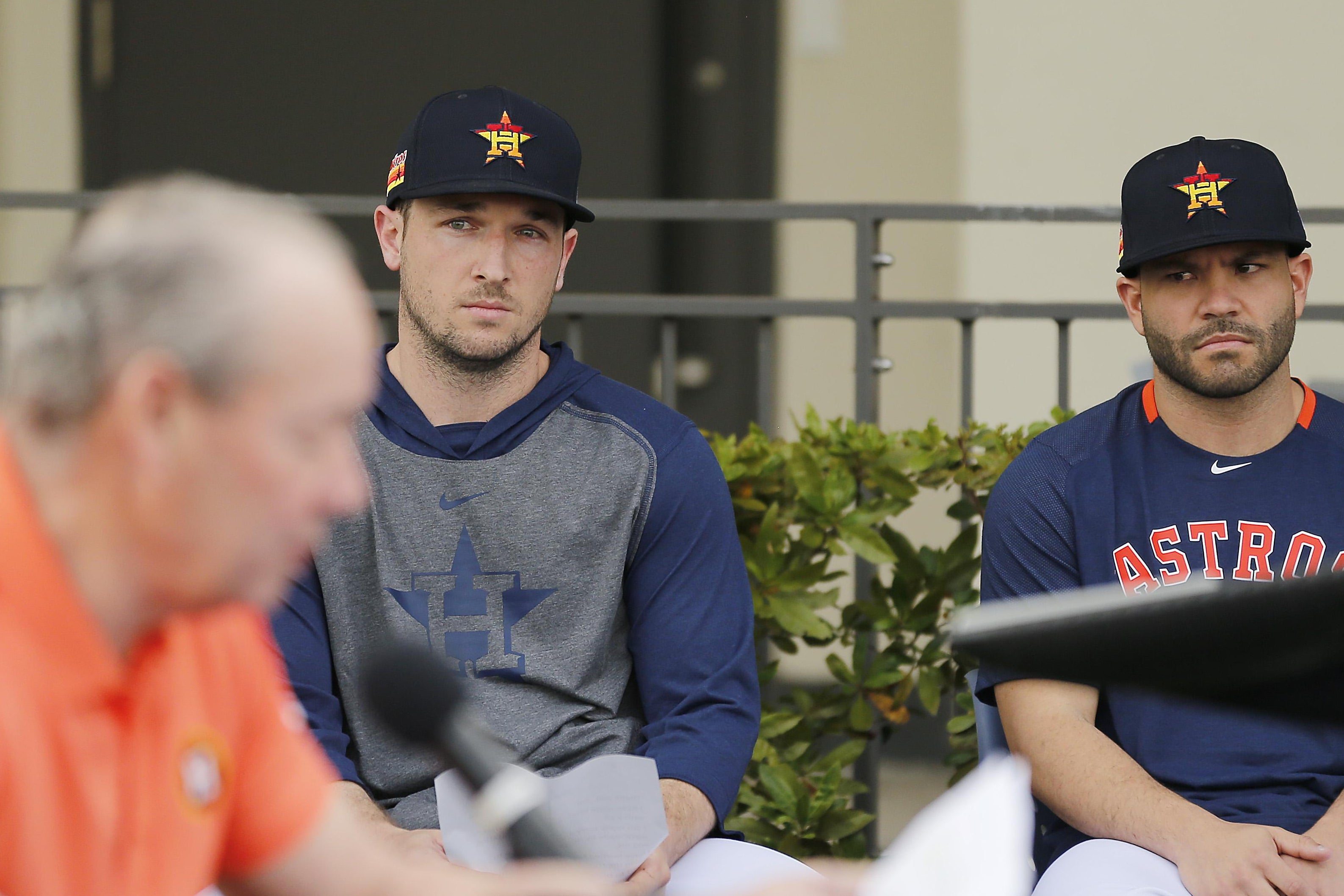 Alex Bregman and Jose Altuve, in warm ups and baseball hats, look on somewhat skeptically as Jim Crane reads seated at a table in front of a microphone in the foreground.