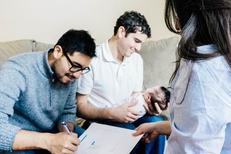 One man holds a baby while another sitting next to him signs paperwork.
