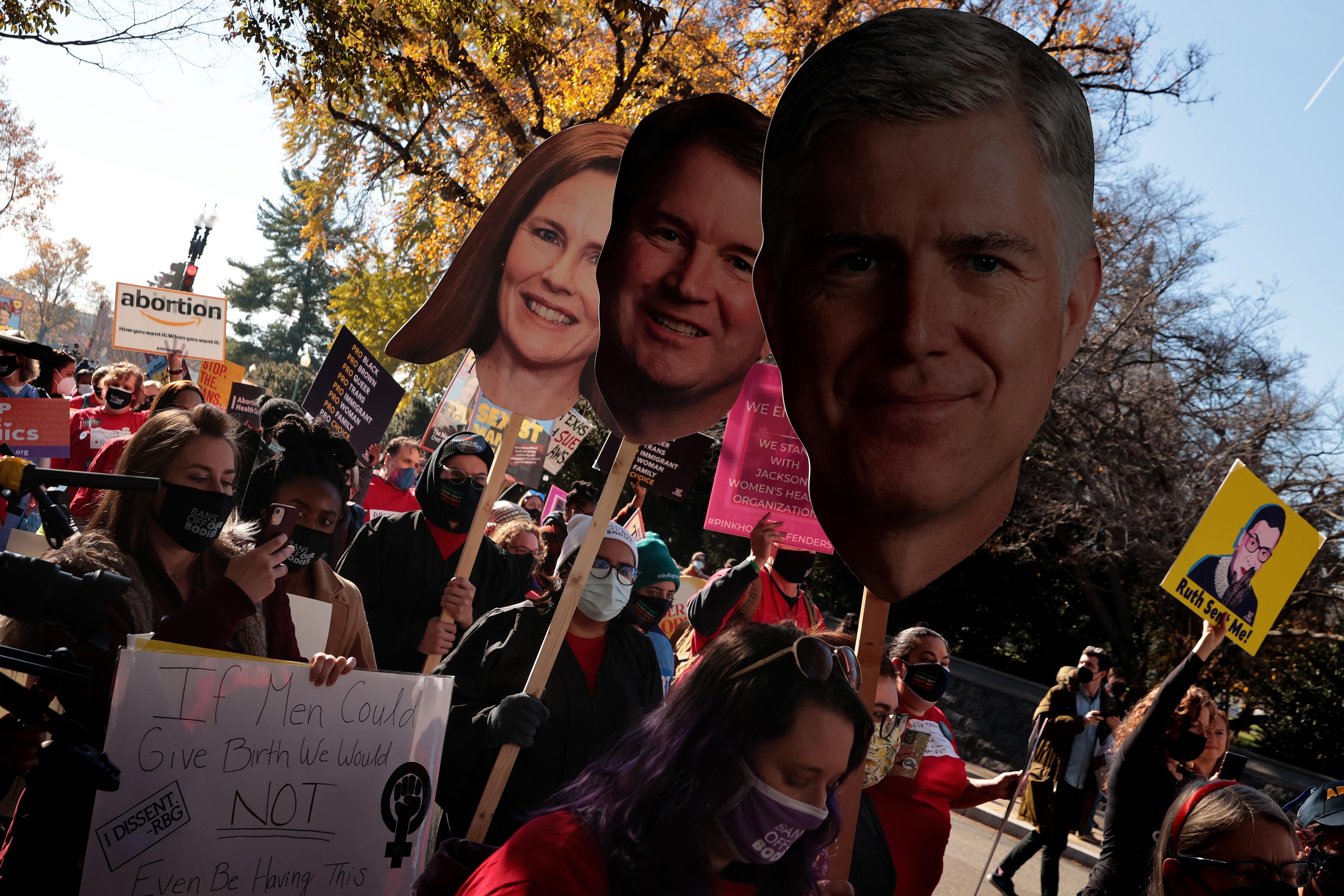 People march with protest signs and large photographic cutouts of Amy Coney Barrett, Brett Kavanaugh, and Neil Gorsuch on a sunny day