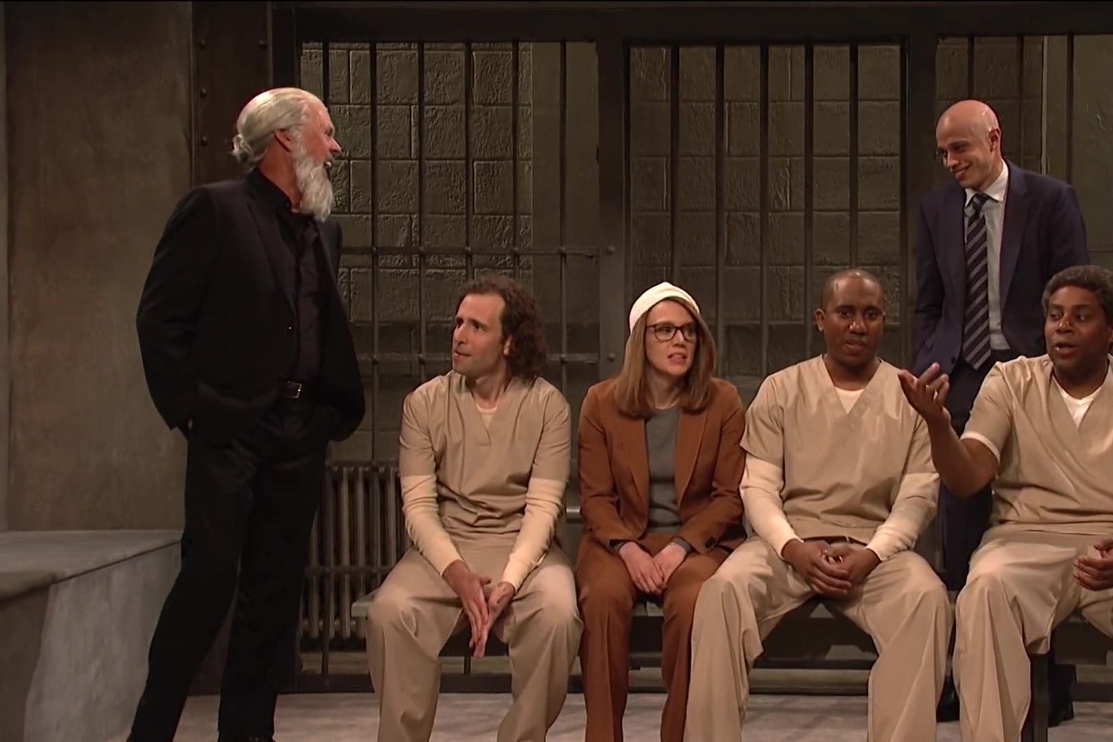 Michael Keaton, as Julian Assange, brags to Kate McKinnon's Lori Loughlin, Pete Davidson's Michael Avenatti, and inmates played by Kyle Mooney, Chris Redd, and Kenan Thompson in a still from SNL.