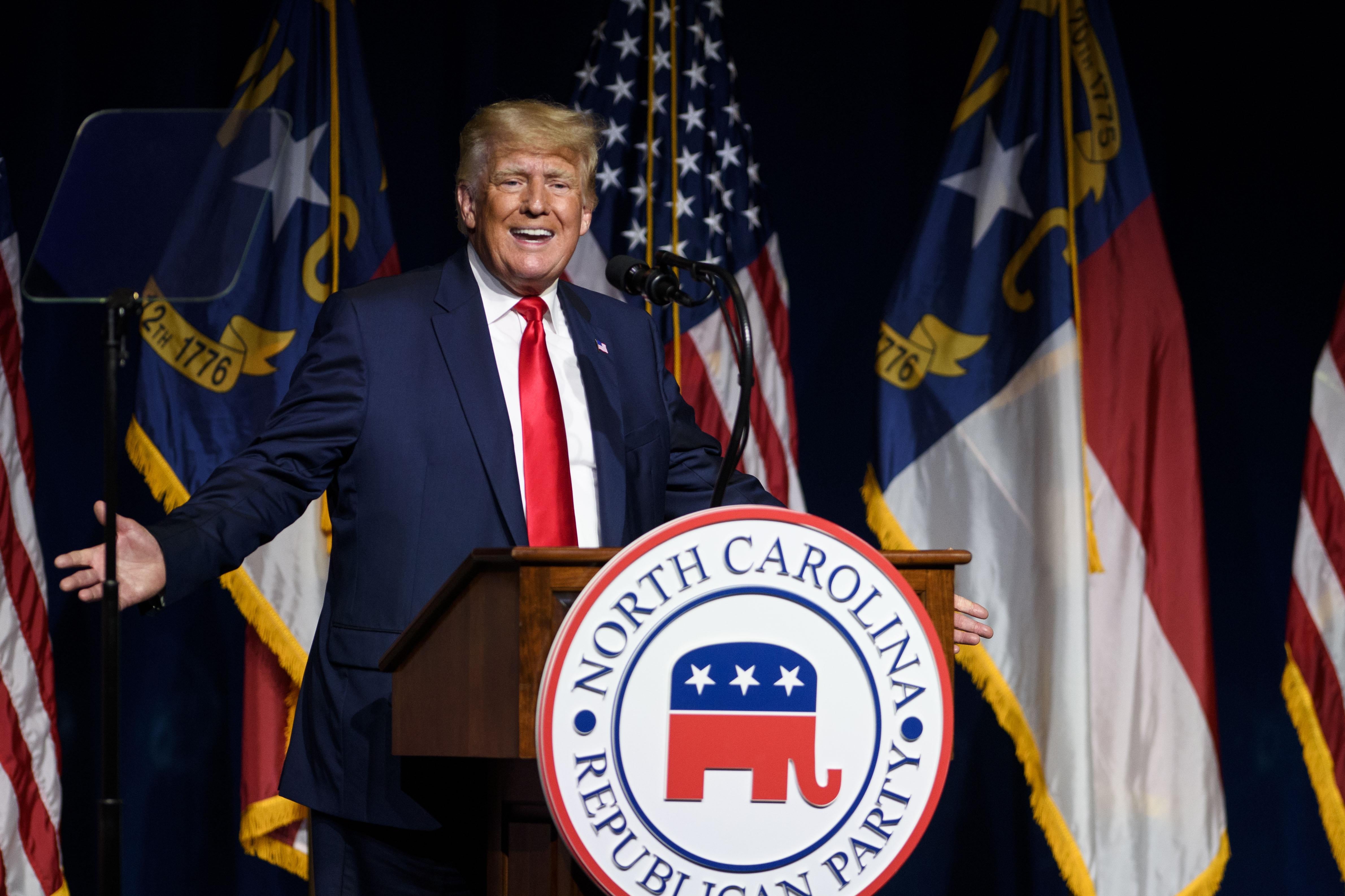 Trump raises both arms at his sides as he speaks at a podium bearing the logo of the North Carolina Republican Party.