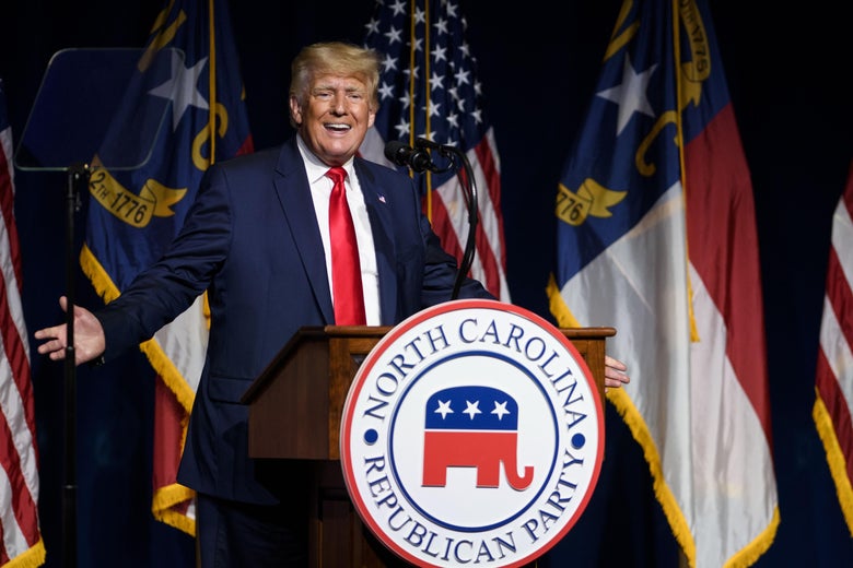 Trump raises both arms at his sides as he speaks at a podium bearing the logo of the North Carolina Republican Party.