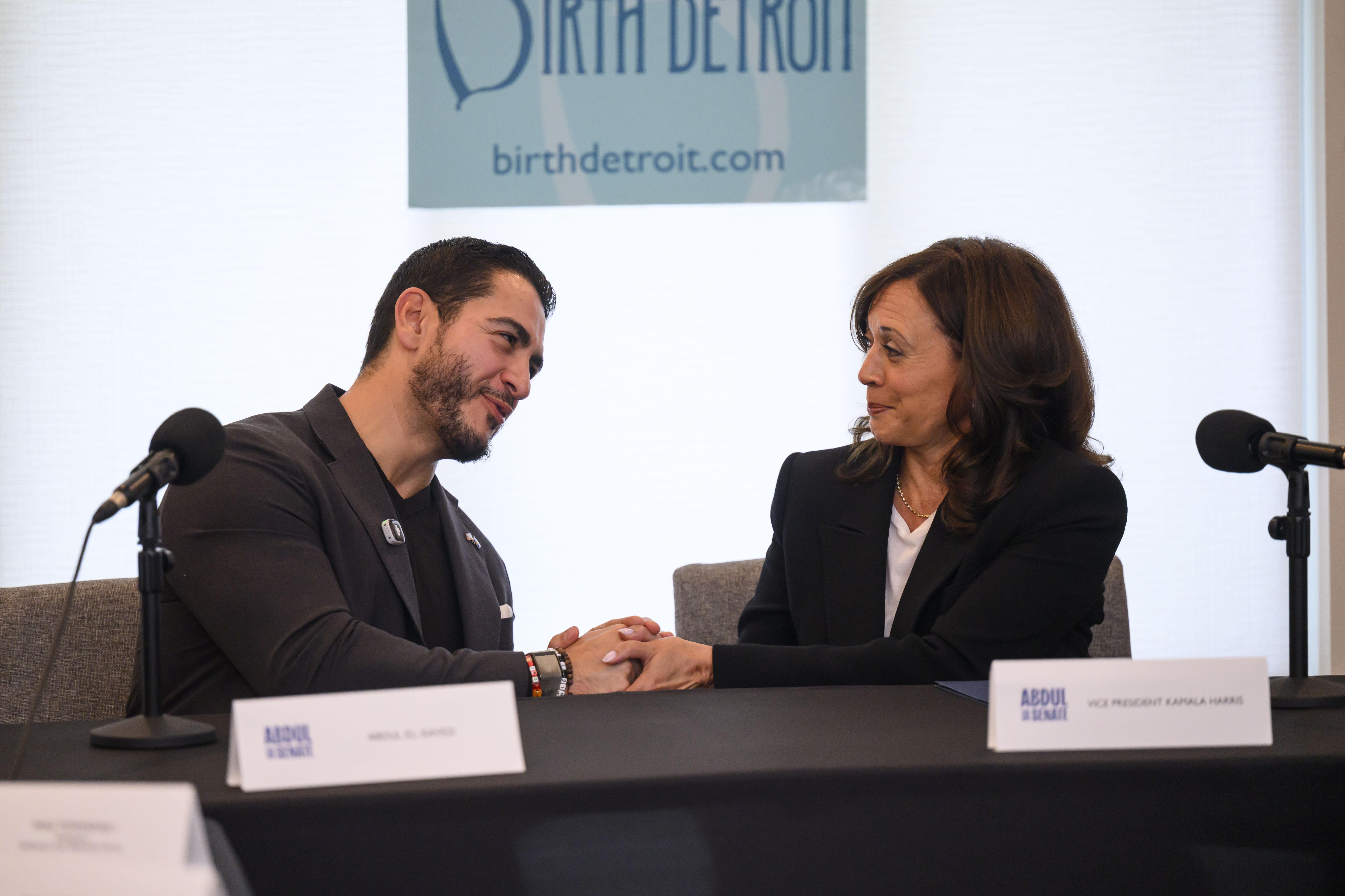 El-Sayed and Harris shake hands while seated next to each other at a table with microphones.