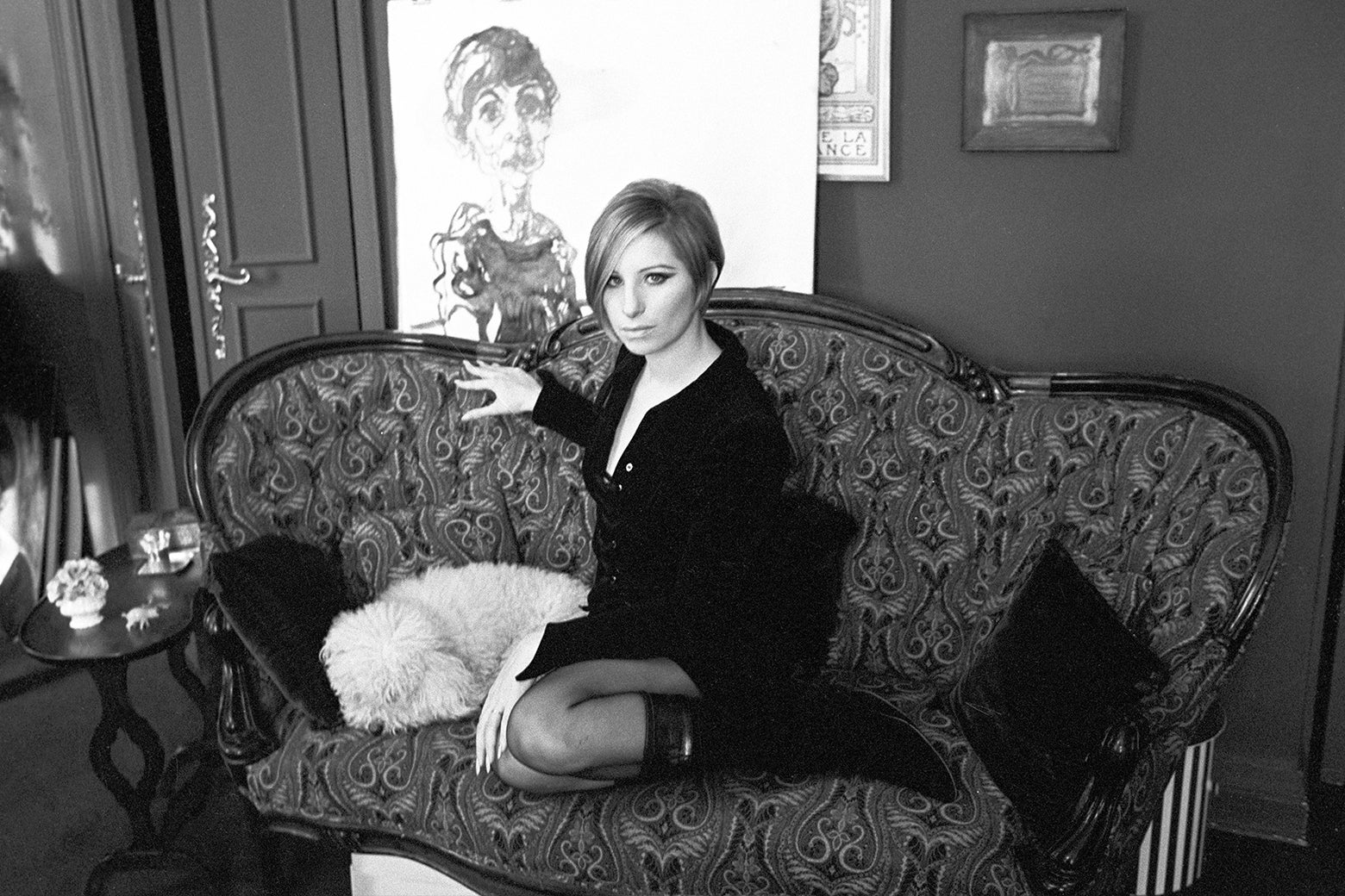 Barbra Streisand, in a 1960s-era bob and knee-high boots and tights, sits on a paisley couch with a white dog.