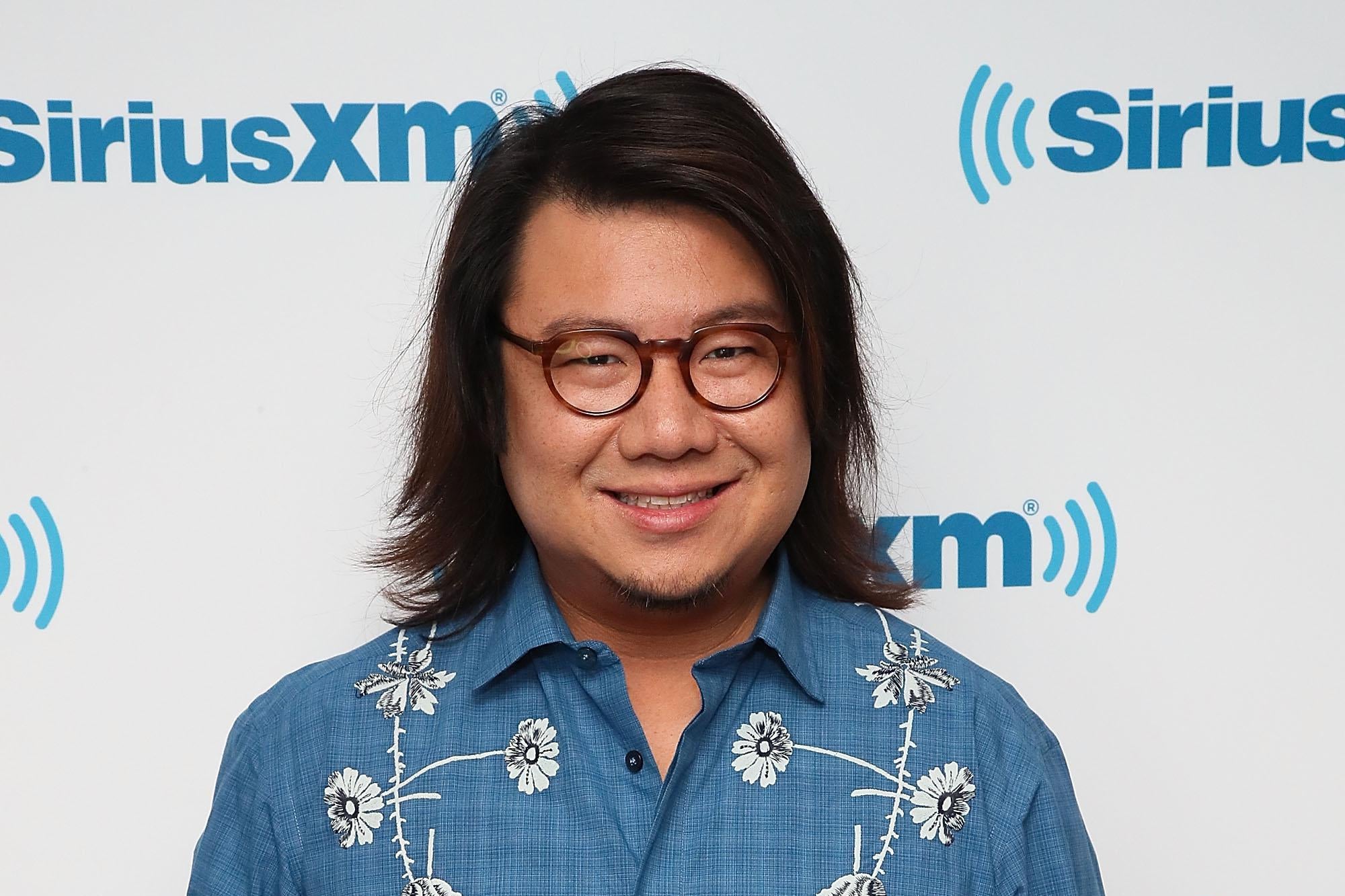 Kevin Kwan, wearing a blue shirt with white flowers and black glasses, stands and smiles at the camera.