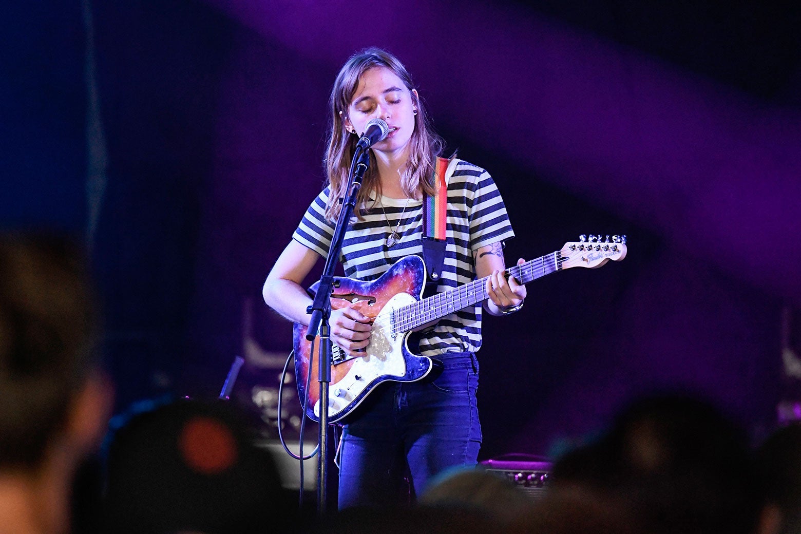A young white woman with shoulder-length hair plays guitar and sings into a microphone.