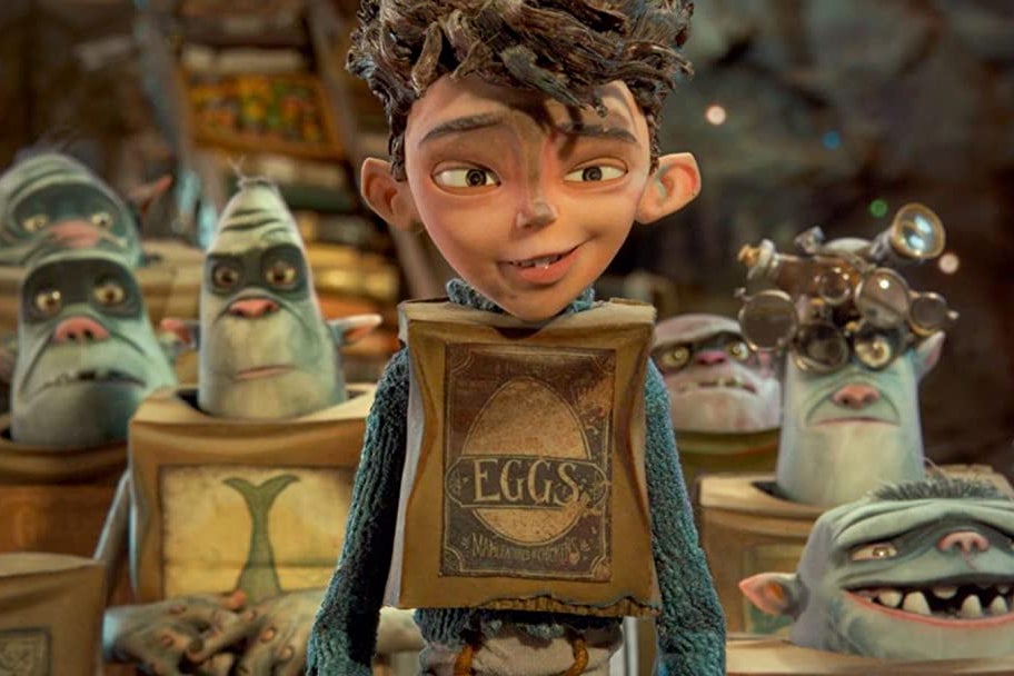 In the style of stop-motion animation: a dark haired boy with a shirt made out of a cardboard box smiles as he stands in front of a group of blue trolls behind him (also dressed with boxes as clothing). 