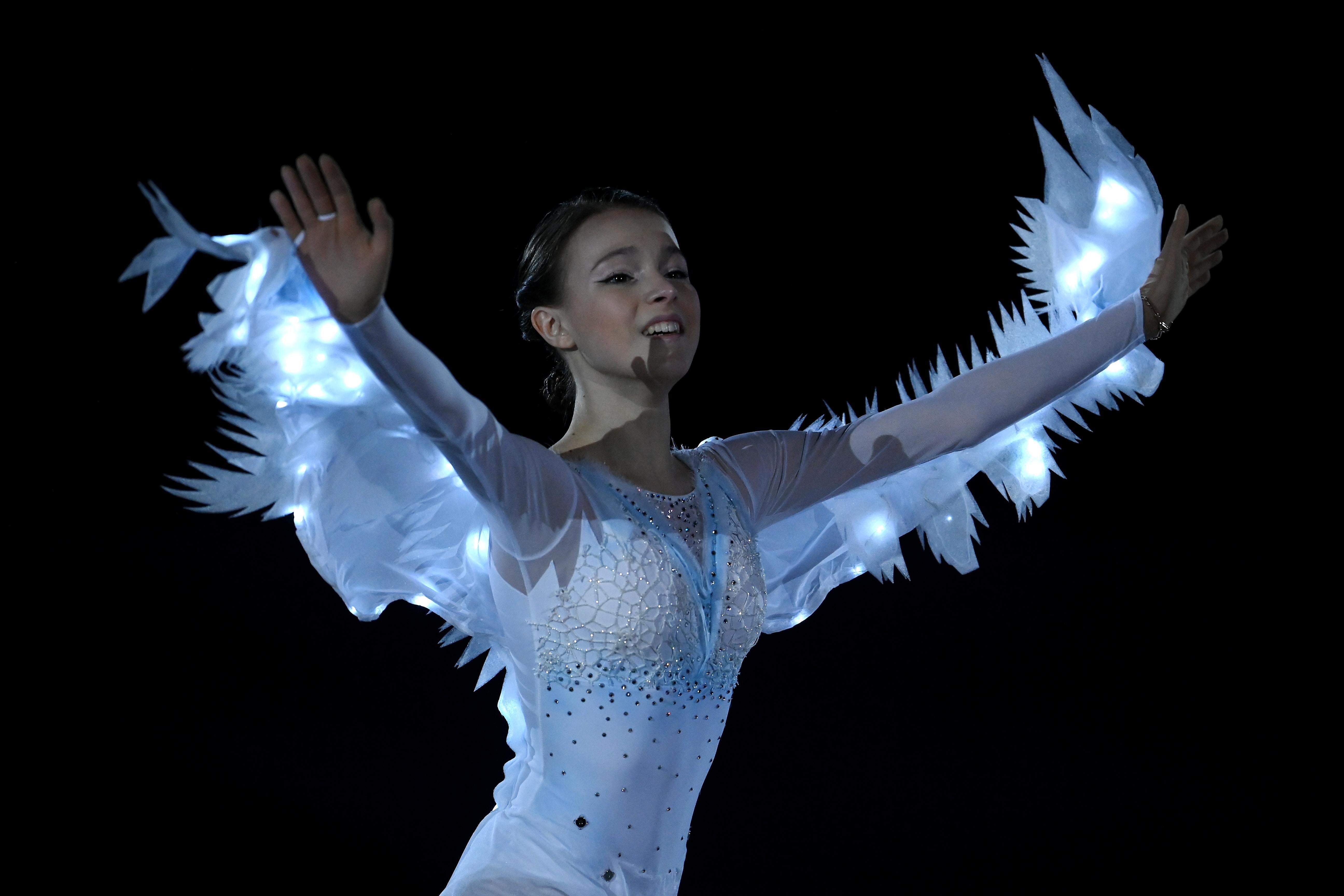 Shcherbakova in a glowing costume with arms featuring feathers, outstretched upward in front of her