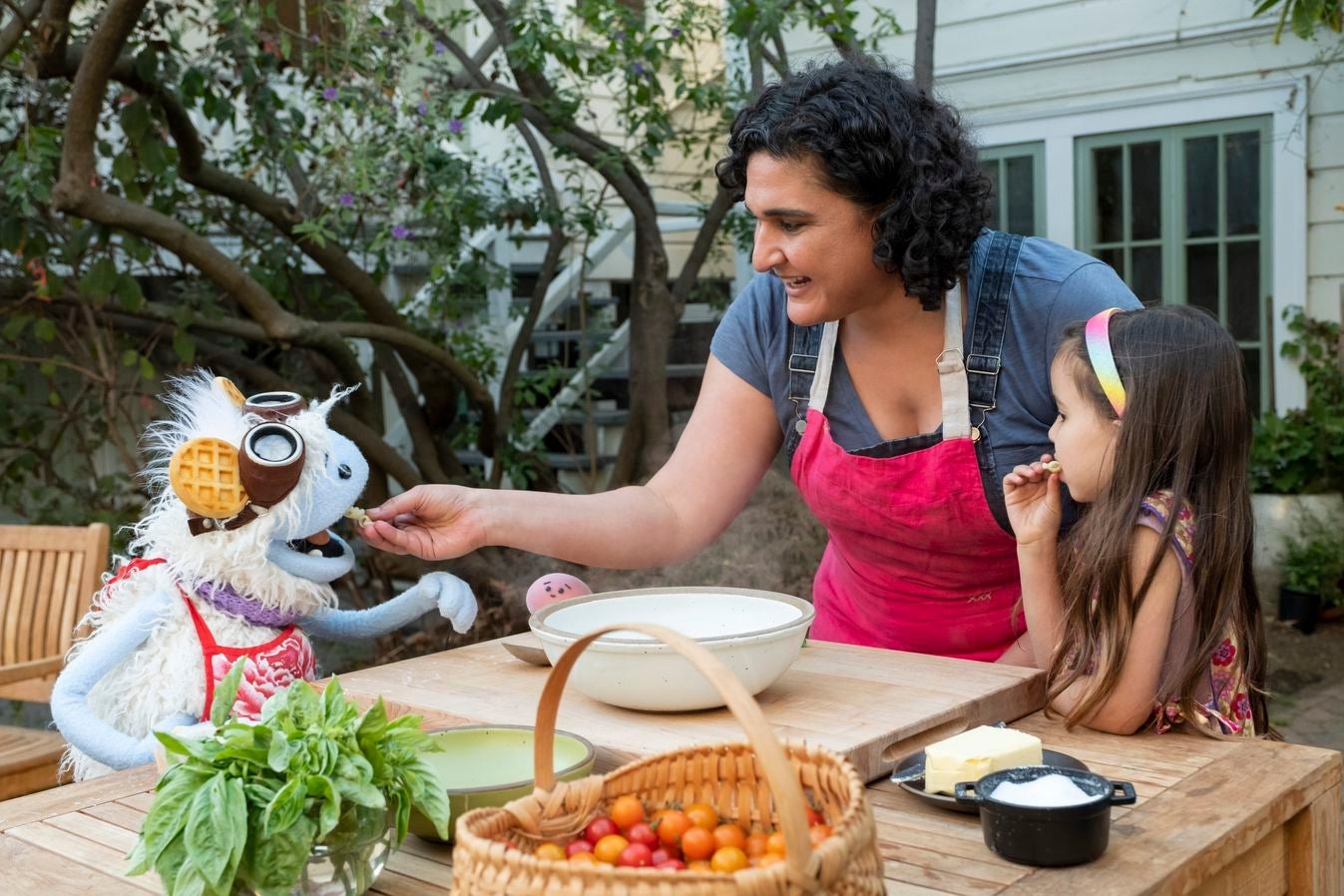 Nosrat feeds Waffles the puppet at an outdoor table as Mochi and a human child look on